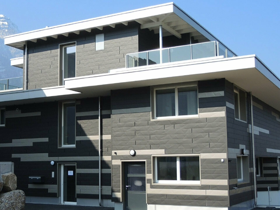 Edificio moderno in svizzera con montagne sullo sfondo con una nuova facciata in Doga.X di PREFA, i pannelli di alluminio sono stati montati in due colori diversi, si sviluppa cosi un designo della facciata individuale