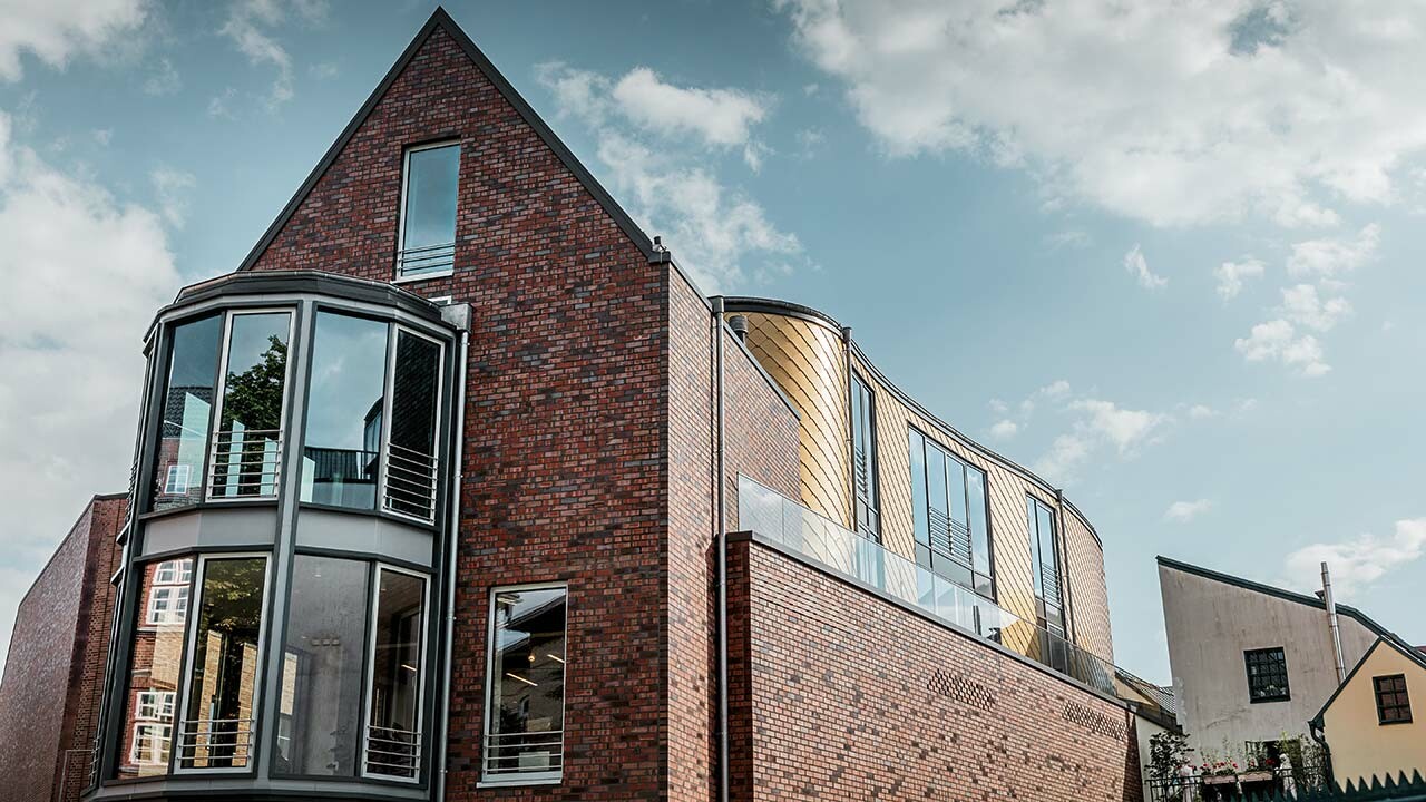 nuovo negozio di calzature Schüttfort ad Amburgo-Bergedorf con una facciata classica in mattoni e una parete curva, rivestito con la scaglia in alluminio di PREFA, il tetto a due falde é rivestito con alluminio grigio, tante vetrate, leggermente nuvoloso