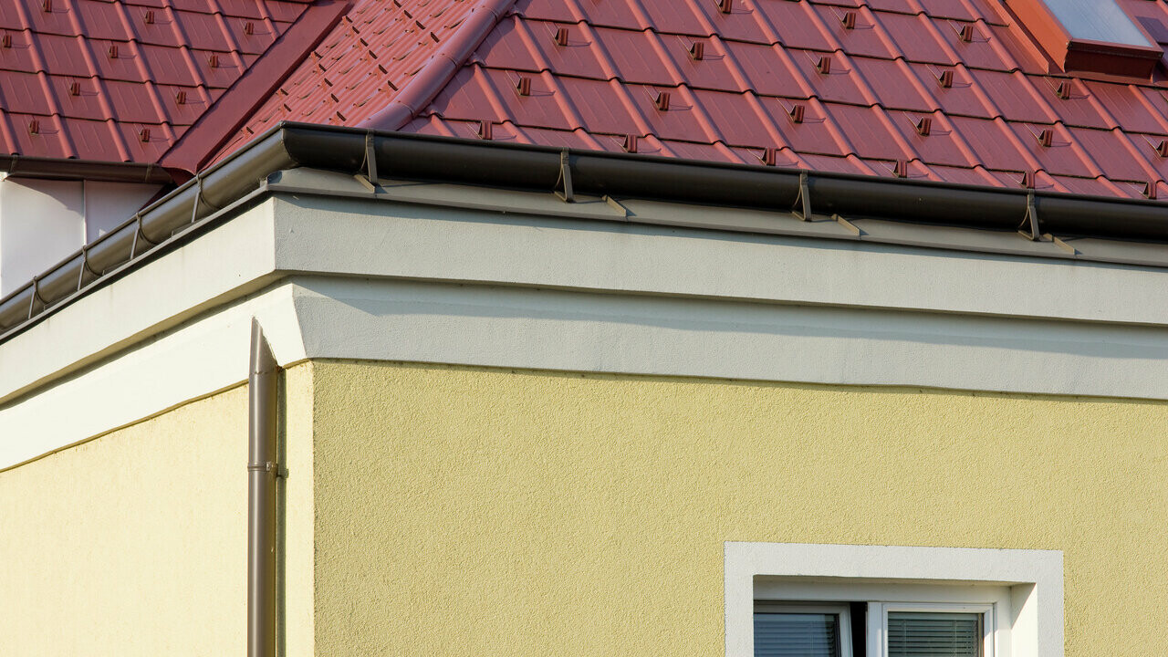 Angolo della casa con facciata gialla intonacata e grondaia PREFA marrone (grondaia cornicione) con tetto a pannelli PREFA rosso