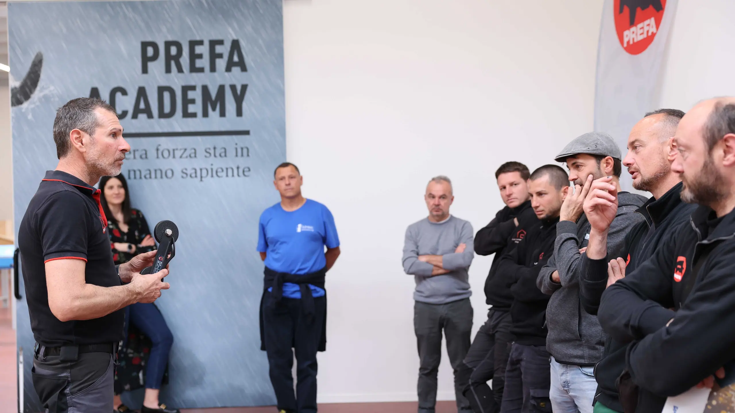 Ein Trainer der PREFA Academy in Italien erklärt einer Gruppe von Handwerkern den Umgang mit PREFA Produkten und Zubehörteilen. Die Teilnehmer hören aufmerksam zu, während sie praktische Anweisungen zur Montage und Verarbeitung von PREFA-Produkten erhalten. Im Hintergrund ist ein PREFA Academy-Banner zu sehen.