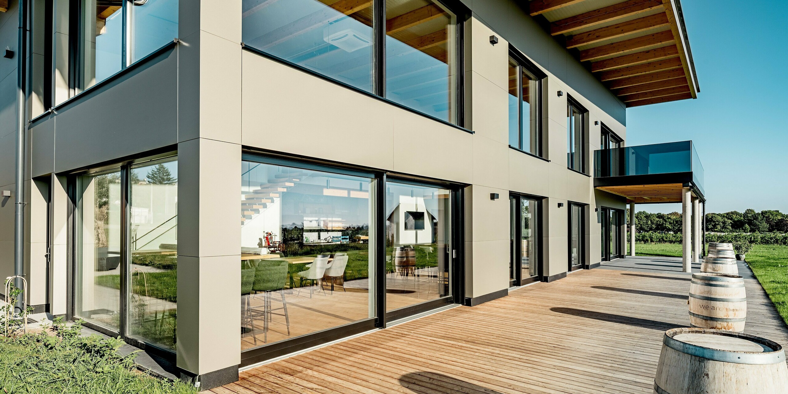 Seitliche Ansicht des Weinguts Nigl in Perchtoldsdorf mit großzügiger Glasfassade und moderner PREFA Aluminiumverkleidung in P.10 Prefabronze. Die filigrane Linienführung der Aluminium Verbundplatten verleiht dem Gebäude eine elegante Struktur, während bodentiefe Fenster viel Licht ins Innere lassen. Die gläserne Balkonbrüstung, das auskragende Holzdach und die sichtbare Treppe im Inneren unterstreichen das hochwertige, offene Architekturkonzept. Auf der Holzterrasse davor stehen Weinfässer als dekoratives Element.