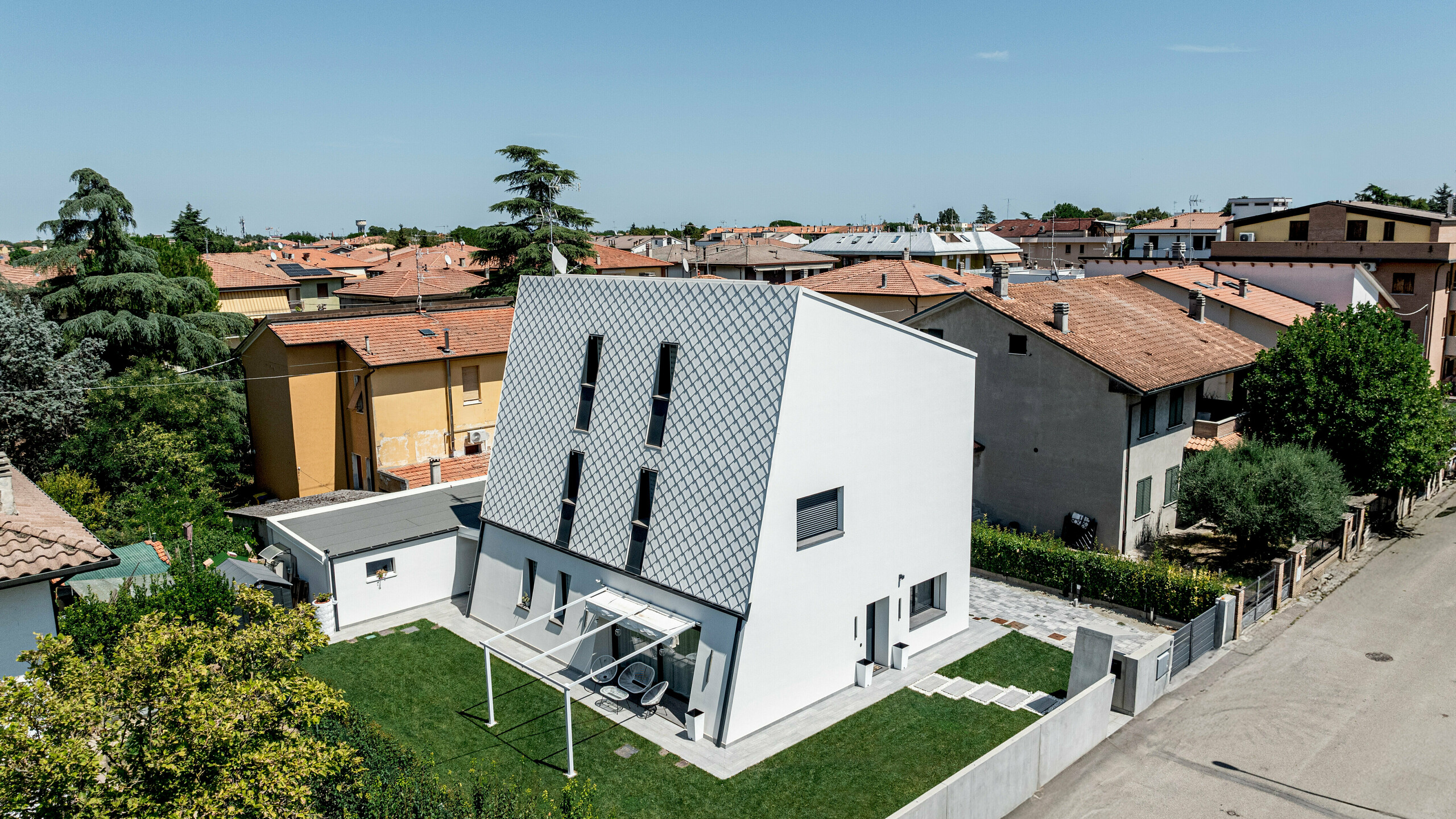 Schräg von oben aufgenommene Ansicht eines modernen Einfamilienhauses in Forlì, Italien, mit markanter, geneigter Hauptfassade aus PREFA Wandraute 29 × 29 in P.10 Reinweiß. Das präzise, diagonal verlaufende Rautenmuster aus Aluminium hebt sich deutlich von den umliegenden Ziegeldächern ab und wird zum architektonischen Blickfang. Klare Linien, schmale Fenster und der gepflegte Garten betonen den zeitgemäßen Charakter. Die PREFA Aluminiumfassade überzeugt durch Langlebigkeit, Witterungsbeständigkeit und eine dauerhaft hochwertige Optik.