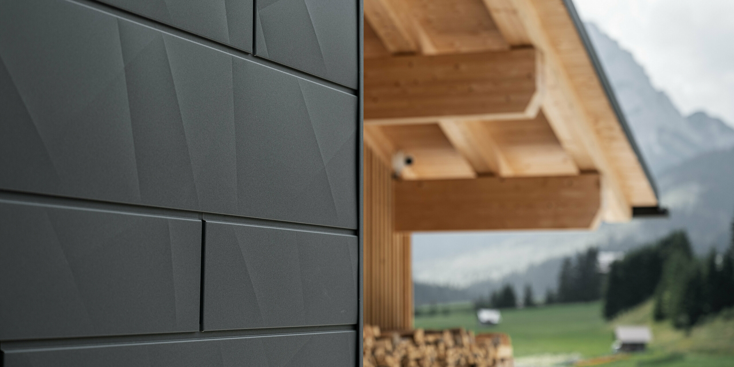 Primo piano dei pannelli di rivestimento Doga.X in P.10 nero di PREFA sulla Casa Hoffe a Sappada, Italia. Il sistema di facciata in alluminio è sapientemente integrato in un design moderno e astratto, che crea un piacevole contrasto con gli elementi in legno a vista della capriata. L'innovativa soluzione per facciate si integra perfettamente nell'ambiente alpino e rappresenta il connubio tra funzionalità, resistenza e cultura edilizia estetica.