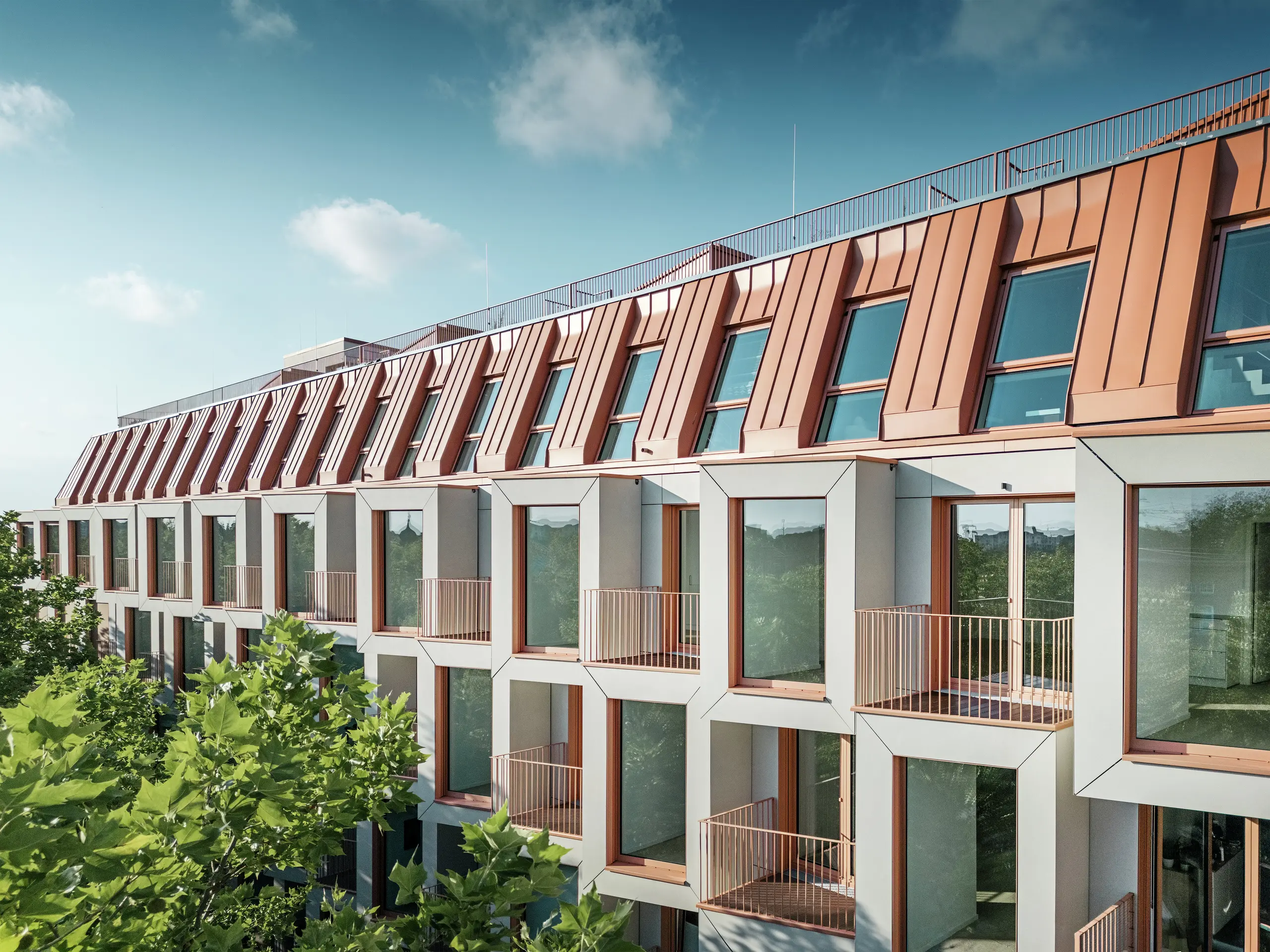 Vista della facciata del progetto residenziale Van B a Monaco di Baviera con il suggestivo tetto in alluminio PREFALZ nel colore speciale IGP Orange HDP di PREFA.  La moderna copertura del tetto combina eleganza estetica e precisione tecnica e offre una protezione duratura dalle intemperie. Le campate dal design cubico con generose superfici finestrate e balconi filigranati creano un'armoniosa interazione di luce, spazio e funzionalità, ideale per sofisticati concetti abitativi in un ambiente urbano.