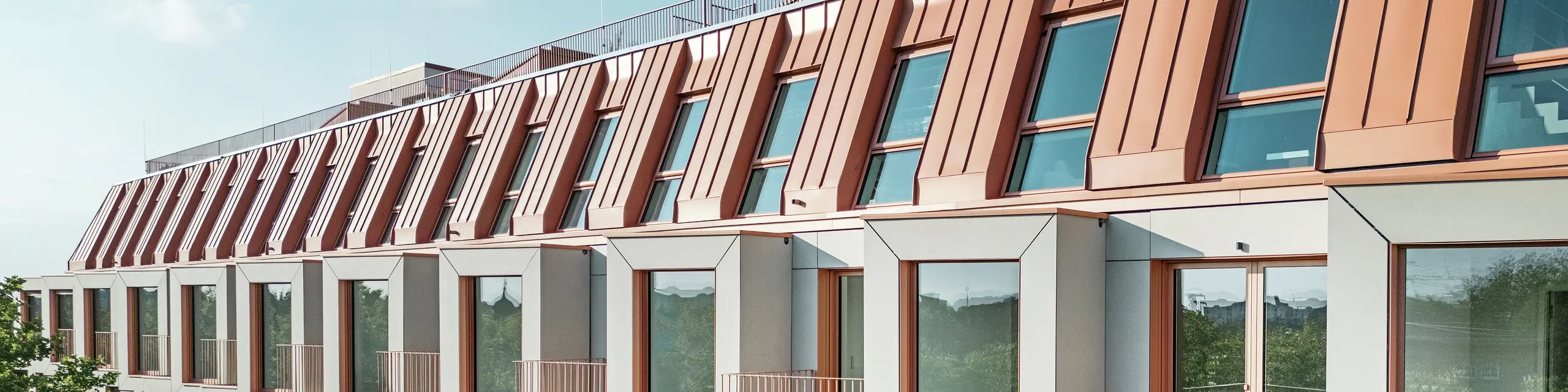 Vista della facciata del progetto residenziale Van B a Monaco di Baviera con il suggestivo tetto in alluminio PREFALZ nel colore speciale IGP Orange HDP di PREFA.  La moderna copertura del tetto combina eleganza estetica e precisione tecnica e offre una protezione duratura dalle intemperie. Le campate dal design cubico con generose superfici finestrate e balconi filigranati creano un'armoniosa interazione di luce, spazio e funzionalità, ideale per sofisticati concetti abitativi in un ambiente urbano.