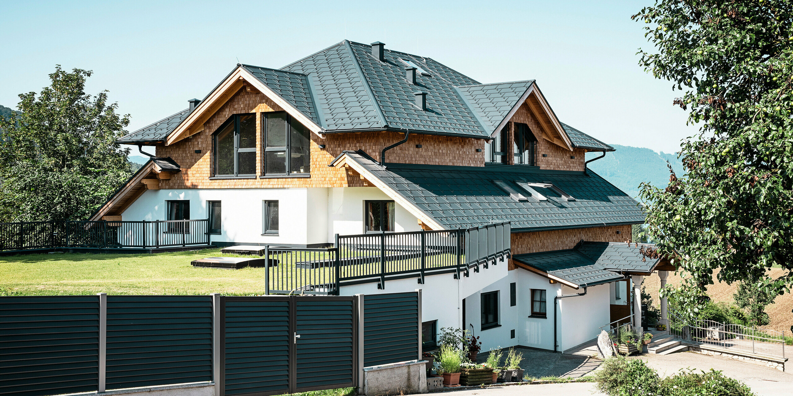 Seitenansicht des Café-Restaurants Zenz'n Stub'n in Schörfling am Attersee mit modernem Aluminiumdach aus PREFA Dachplatten R.16 in der hochwertigen Beschichtung P.10 Anthrazit. Das langlebige PREFA Dach vereint Ästhetik und Funktionalität und bietet optimalen Schutz. Der Zugang zum Garten und die harmonische Einbettung des Gebäudes in die natürliche Umgebung unterstreichen das zeitlose Design.