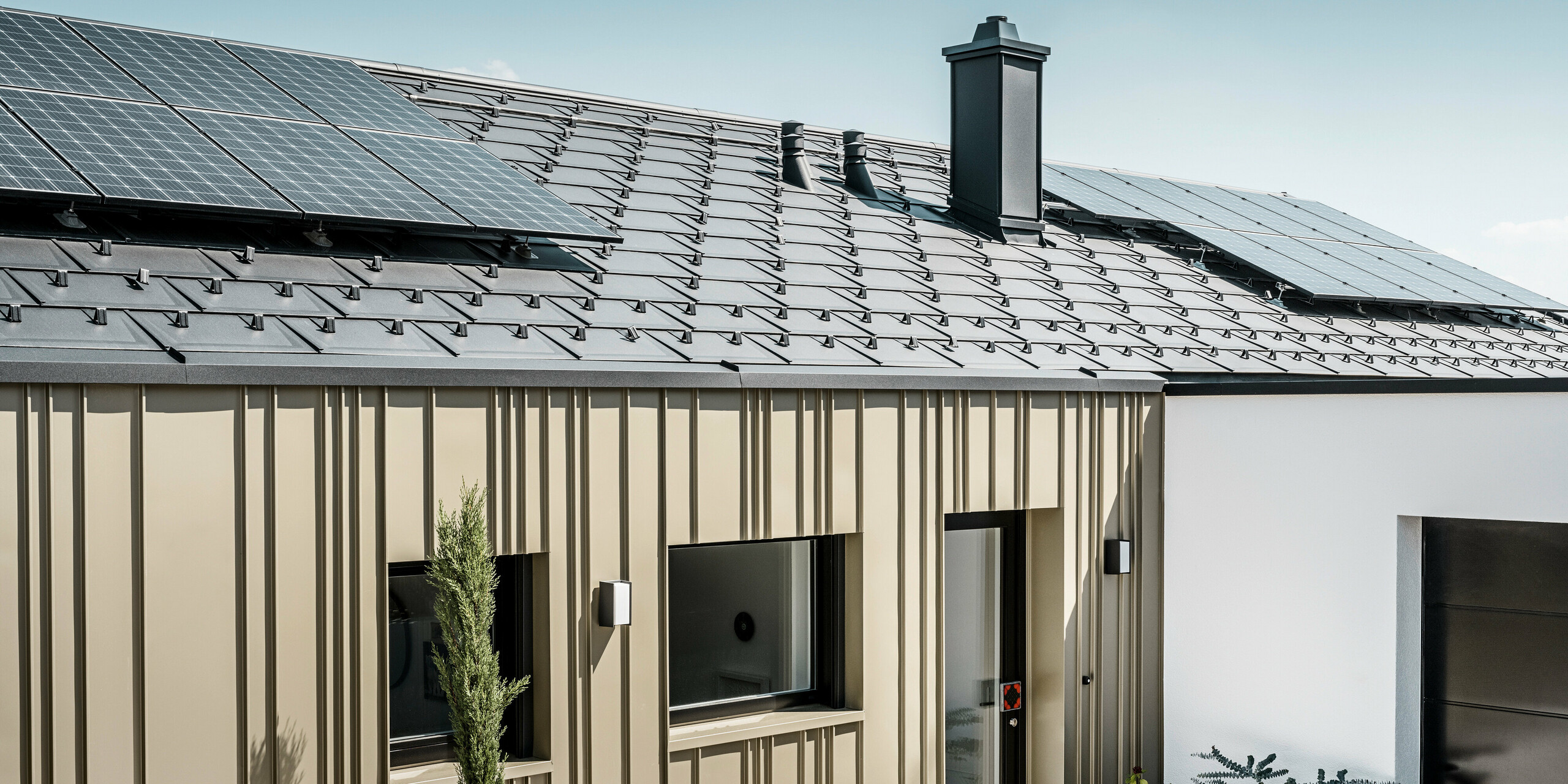 Energieeffizientes Einfamilienhaus mit einem langlebigen PREFA Aluminiumdach in Anthrazit, ausgestattet mit Photovoltaikmodulen. Die zukunftsweisende Wohnlösung wird zudem von einer robusten und widerstandsfähigen PREFA Aluminiumfassade in Lichtbronze betont. Die Fassade weist eine vertikale Strukturierung auf, die durch die anthrazitfarbene PREFA Dachplatte R.16 ergänzt wird. Diese Kombination bietet nicht nur ästhetische Schönheit, sondern auch Nachhaltigkeit und Energieunabhängigkeit in der modernen Architektur. Ergänzt wird der nachhaltige Bau durch eine PV-Anlage auf dem Dach.