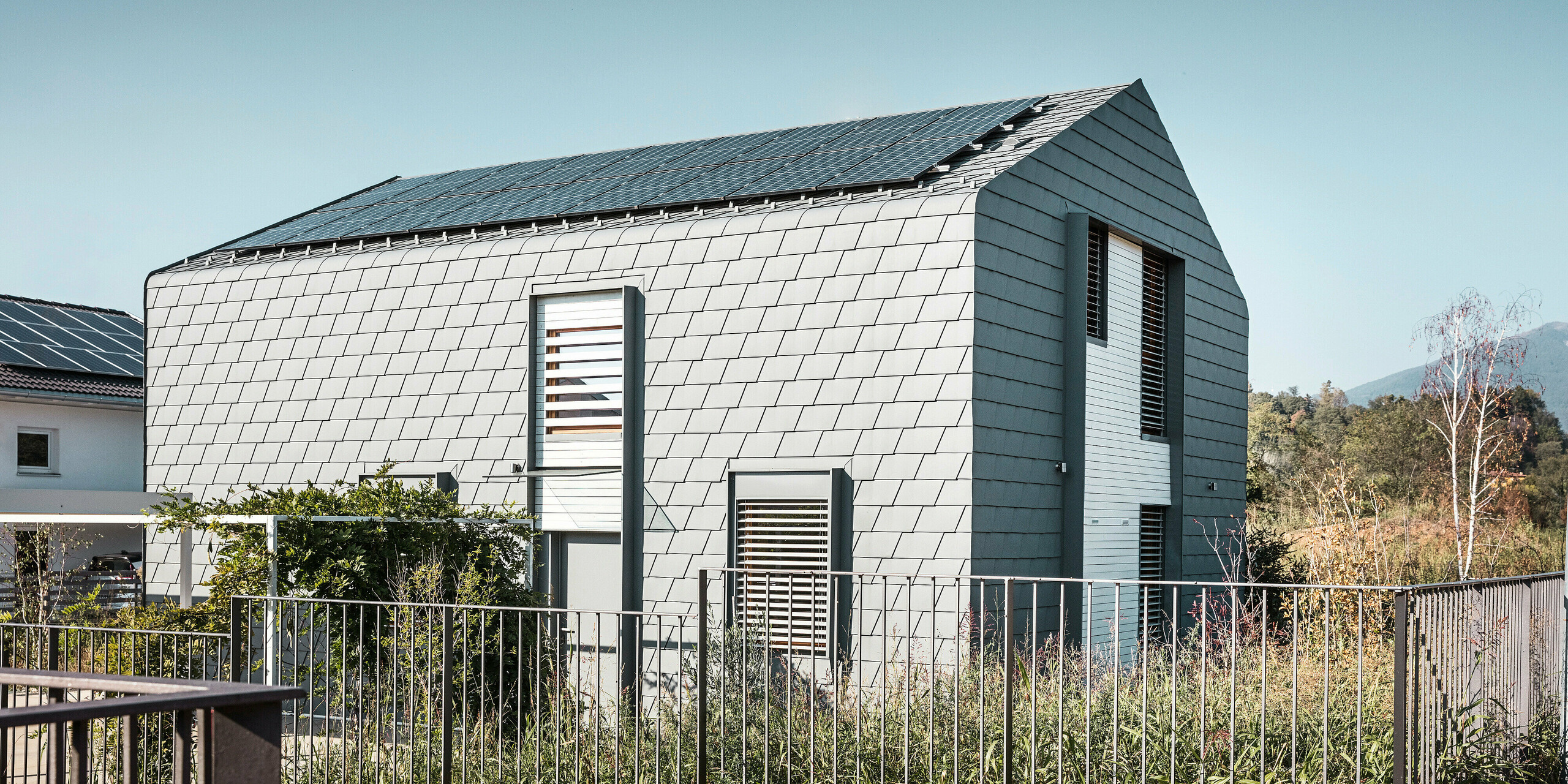 Seitliche Ansicht auf Casa Longaretti. Das moderne Gebäude wird fast vollständig von PREFA Dachschindeln in P.10 Hellgrau ummantelt. Unterbrochen wird die holistische Aluminiumhülle nur von Fenstern und Türen. Die robusten Schindeln mit ihrer schuppenartigen Textur unterstreichen die Qualität und Innovationskraft von PREFA Aluminiumprodukten. Das kleinformatige Dach- und Fassadenprodukt steht für Langlebigkeit und Witterungsbeständigkeit sowie eine zeitgemäße Architektur. Das kompakte Einfamilienhaus verfügt über einen kleinen Garten mit zahlreichen Pflanzen. Das Dach wird auf dieser Seite fast gänzlich von einer Photovoltaikanlage eingenommen. 