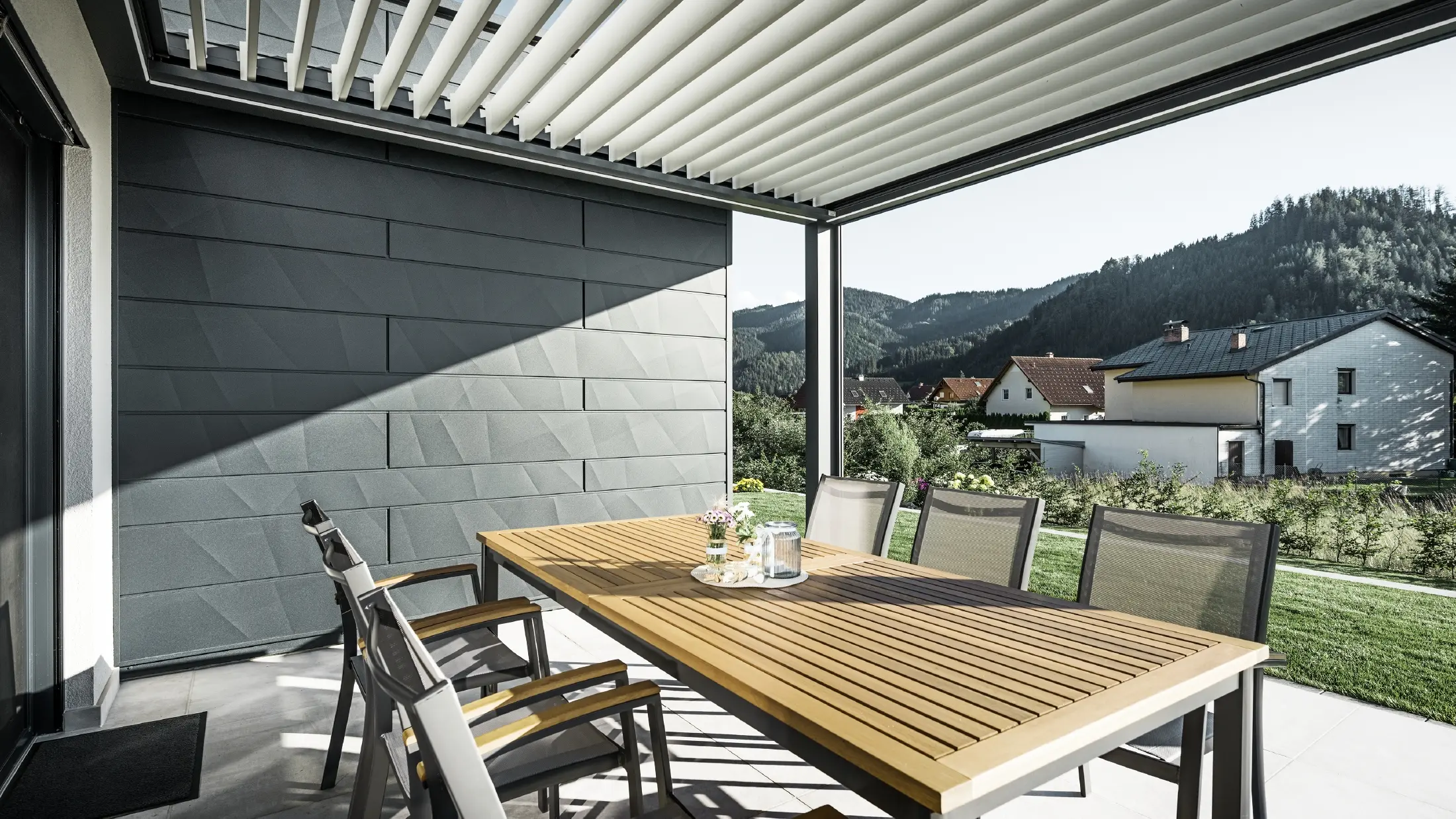 Terrazza coperta da pergolato a lamelle con arredi esterni; parete di fondo rivestita con pannelli per facciata Doga.X PREFA in colore antracite.