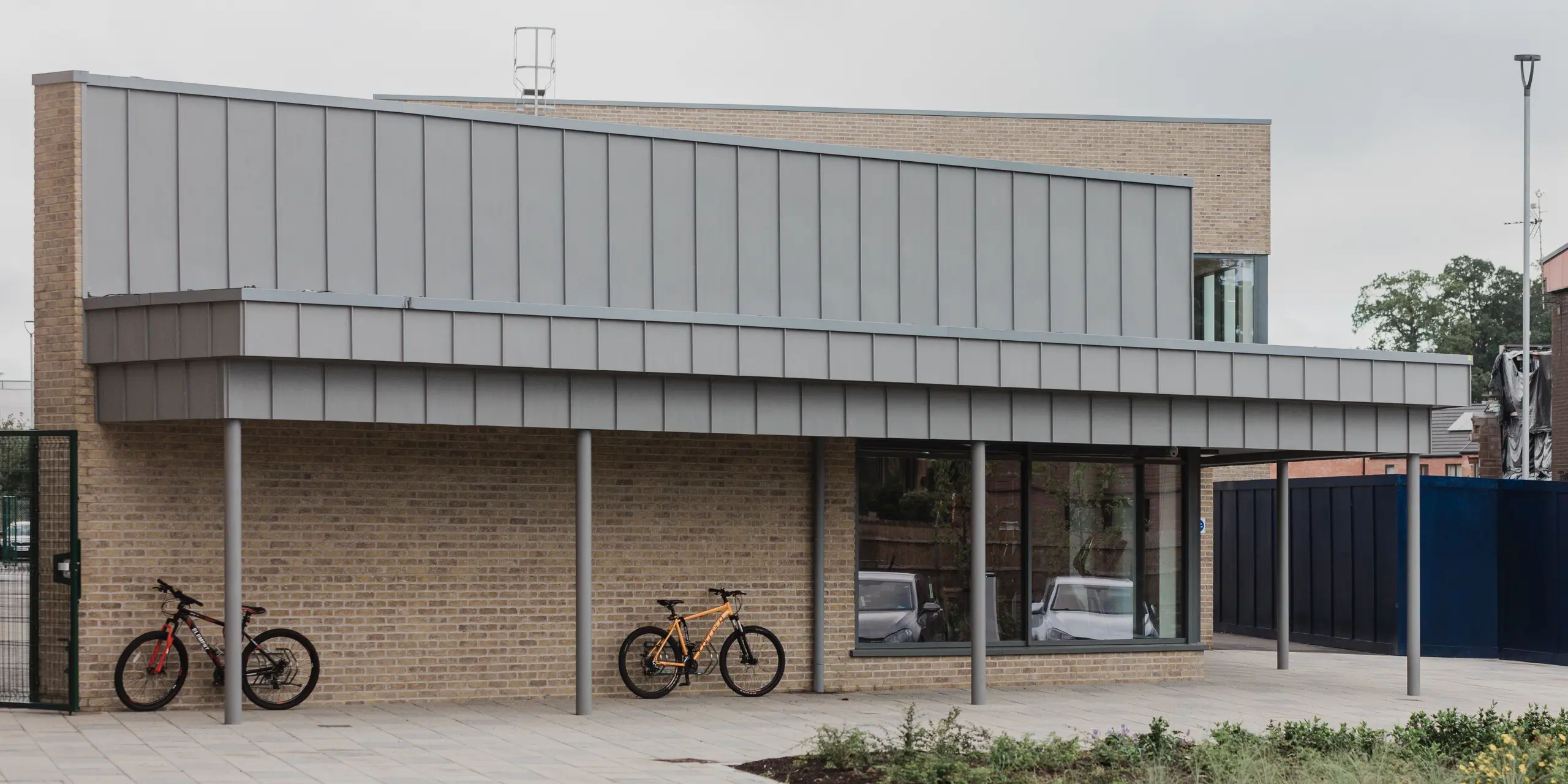 Das Northern Regional College in Ballymena verfügt über einen modernen Fahrradstellplatz mit großzügigem Vordach aus PREFALZ Aluminium in der Farbe Patinagrau. Die vertikal strukturierten Stehfalzbahnen ziehen sich präzise über die Dachkante und bilden einen klaren Blickfang über der Klinkerfassade. Schlanke Stützen, helle Klinkerwände, abgestellte Fahrräder und der offene Vorplatz unterstreichen den nutzerfreundlichen Charakter des Campus. PREFA Aluminium überzeugt durch Langlebigkeit, Witterungsbeständigkeit und eine ruhige, hochwertige Optik im täglichen Betrieb.