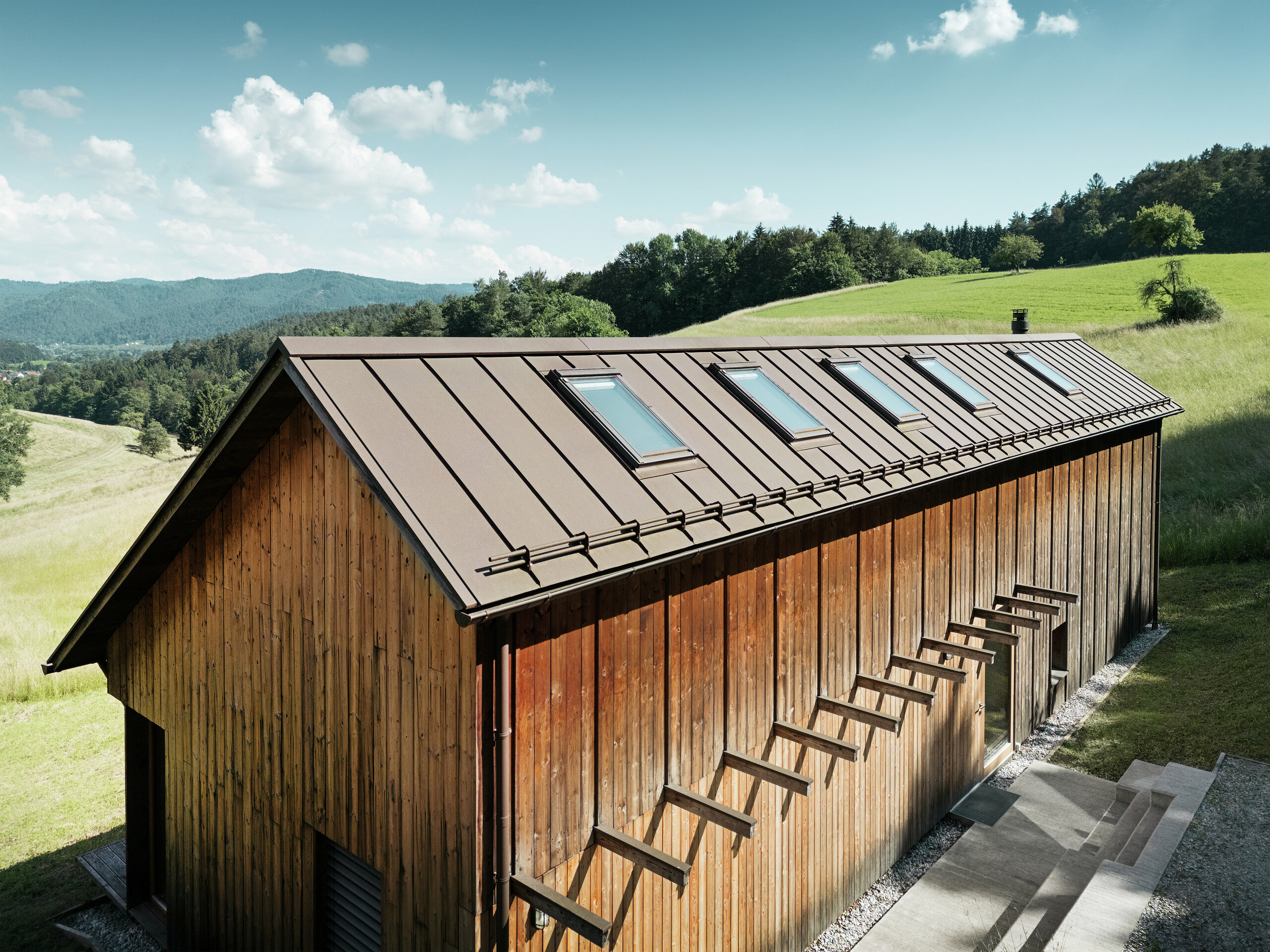 Casa in legno dalla struttura architettonica chiara con tetto in alluminio PREFALZ di alta qualità in P.10 marrone. Il tetto PREFA, che richiede poca manutenzione, convince per le sue linee precise, l'elevata resistenza ai raggi UV e alla corrosione e la perfetta integrazione delle finestre per tetti. La combinazione di una facciata in legno sostenibile e di un tetto doppia aggraffatura durevole si inserisce armoniosamente nell'idilliaco paesaggio collinare. Il tetto a falde asimmetriche è dotato di cinque lucernari posizionati con precisione che forniscono luce all'interno.