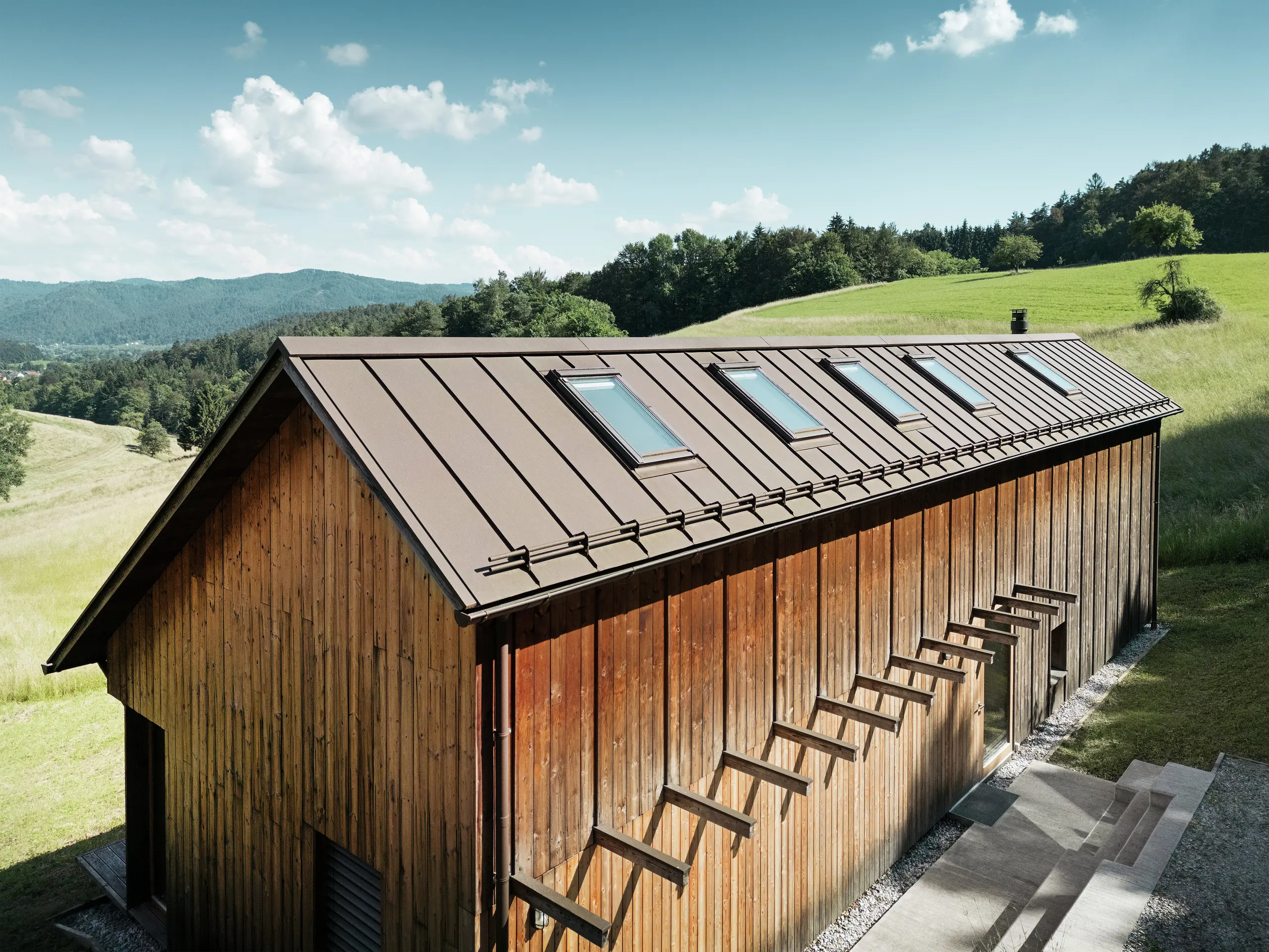 Architektonisch klar strukturiertes Holzhaus mit hochwertigem PREFALZ-Aluminiumdach in der Farbe P.10 Braun. Das pflegeleichte PREFA-Dach besticht durch präzise Linien, hohe UV- und Korrosionsbeständigkeit sowie die perfekte Integration der Dachfenster. Die Kombination aus nachhaltiger Holzfassade und langlebiger Stehfalz-Dachdeckung fügt sich harmonisch in die idyllische Hügellandschaft ein. Das asymmetrische Satteldach verfügt über fünf präzise positionierte Dachfenster, die für natürliches Licht im Inneren sorgen.