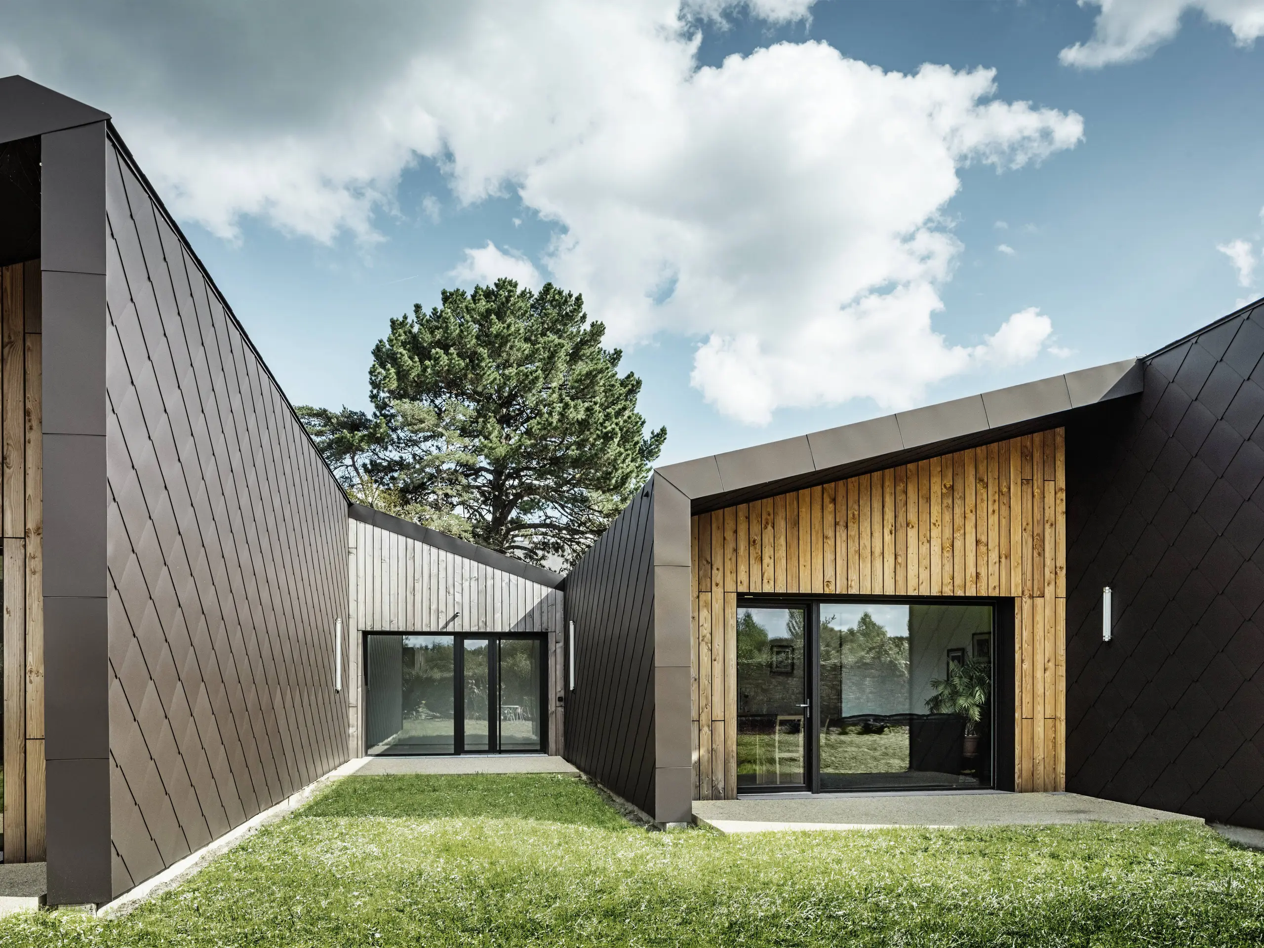 L'immagine mostra un'architettura moderna con una facciata composta da scaglie  44 × 44 PREFA per tetto e facciate nel colore marrone testa di moro. La struttura a diamante del rivestimento in alluminio conferisce all'edificio un aspetto elegante e geometrico. La facciata combina armoniosamente elementi in legno naturale e ampie superfici vetrate, creando un'atmosfera calda e invitante. Il prato verde e il cielo limpido rafforzano l'impressione di modernità e naturalezza dell'architettura.