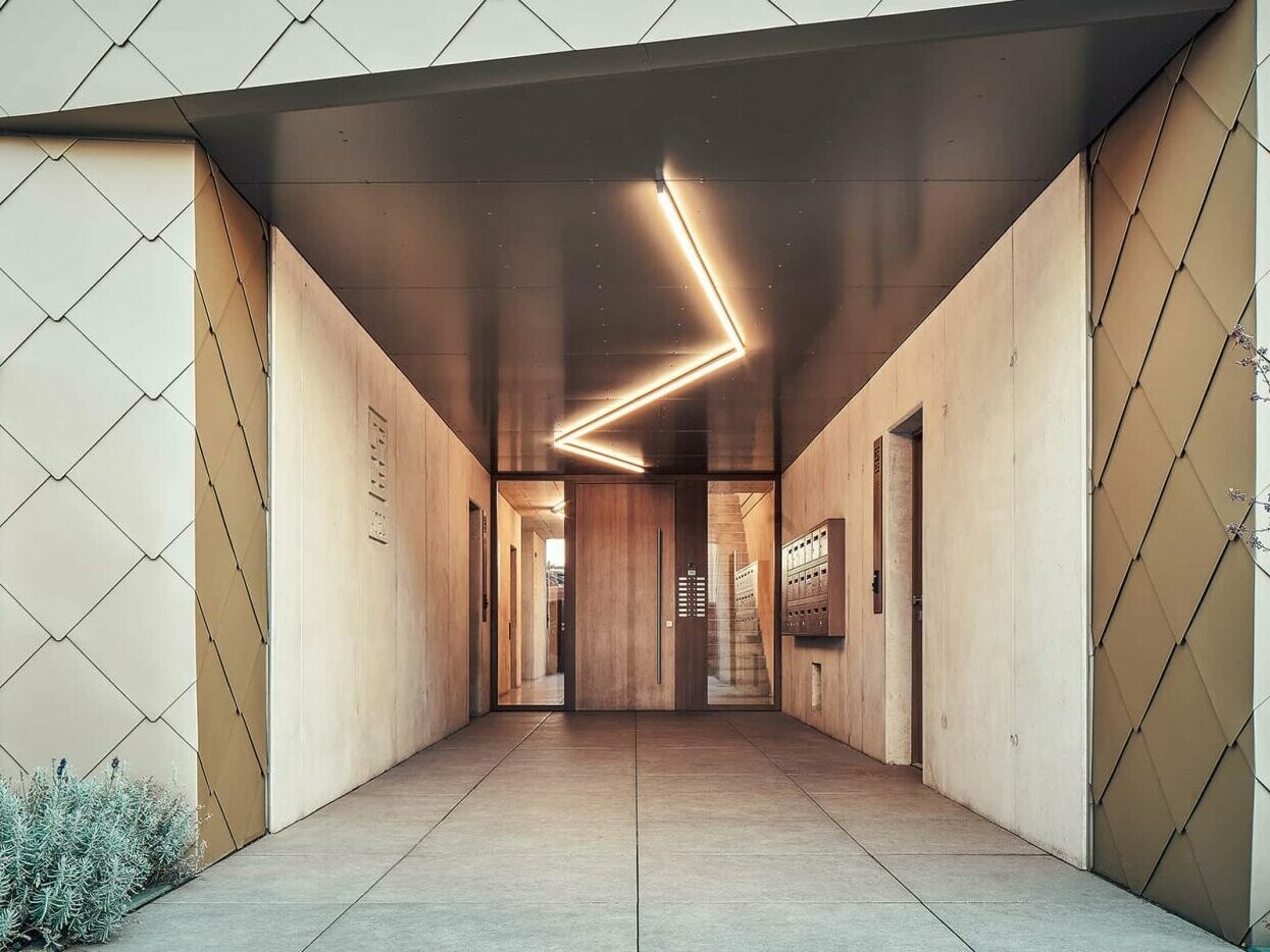 L'immagine mostra il suggestivo ingresso illuminato dell'edificio residenziale con calcestruzzo ed elementi di facciata di PREFA..