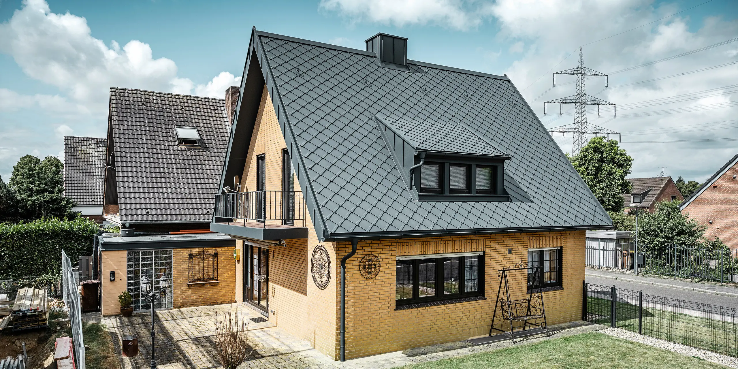 Ein Einfamilienhaus in Tönisvorst mit einem PREFA Dach aus Dachrauten 29×29 in P.10 Anthrazit, das ein herausstechendes Rautenmuster bietet. Schleppgauben, Kamin und der Eingangsbereich sind mit PREFALZ verkleidet, was zusammen mit Ablaufrohr und Hängerinne in P.10 Anthrazit dem Haus einen zeitlosen und eleganten Look verleiht. Das Backsteingebäude mit dem Aluminiumdach harmoniert mit der ruhigen, grünen Umgebung der Siedlung.
