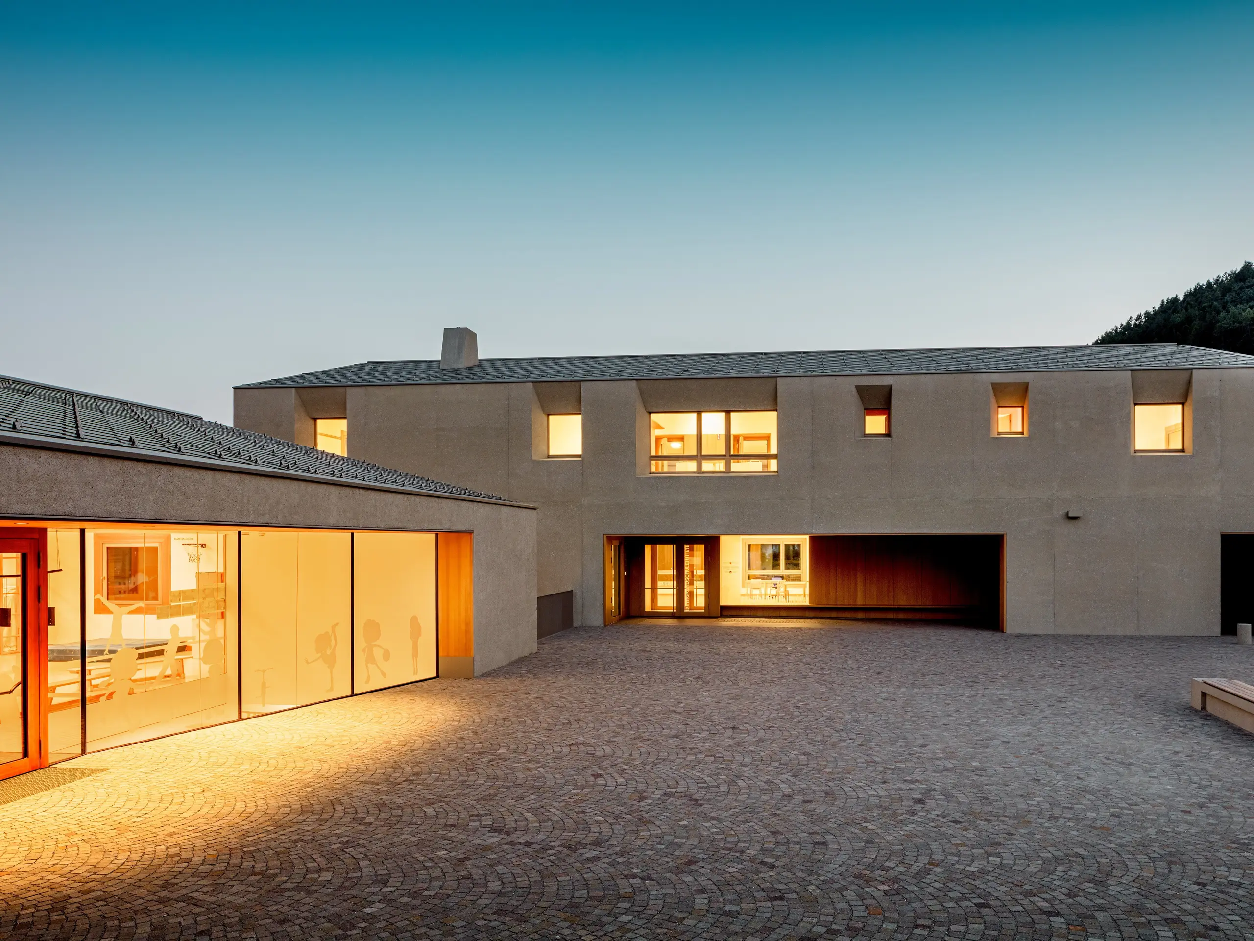 Al crepuscolo, il centro educativo di Riffian risplende di una luce calda sul cortile lastricato della scuola. Le scaglie 44 × 44 PREFA in P.10 bronze coronano la scuola materna e la scuola elementare con una copertura in alluminio dalle linee precise, coniugando un’architettura pulita con una lunga durata resistente alle intemperie e creando un accento elegante e sereno sullo sfondo del versante montuoso altoatesino.