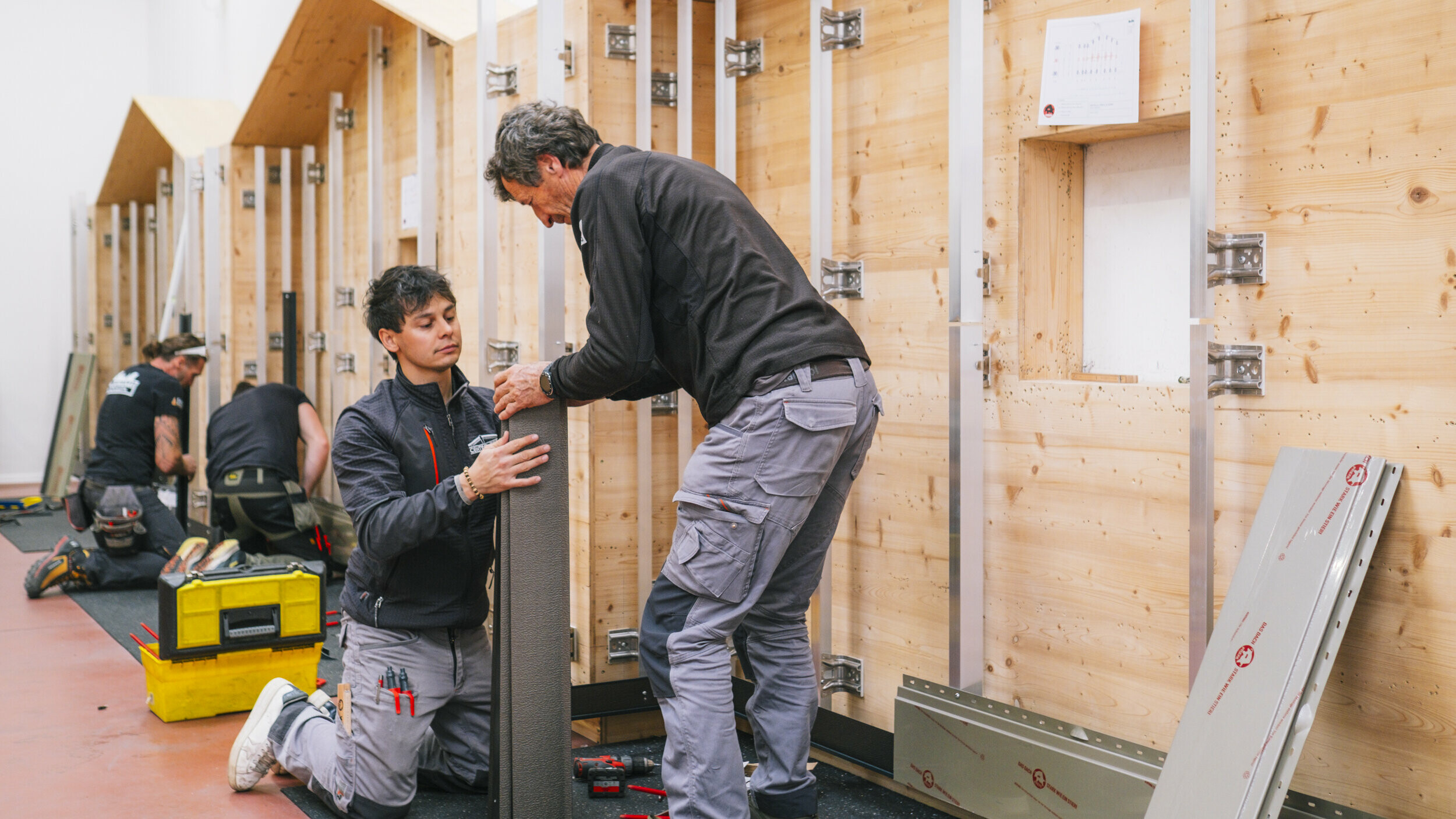 I partecipanti a un corso pratico presso la PREFA Academy Italia lavorano su una parete di prova in legno con sottostruttura in alluminio. Due persone in primo piano montano insieme un elemento di facciata. La scena mostra la formazione artigianale nel campo dei sistemi per facciata e copertura PREFA.