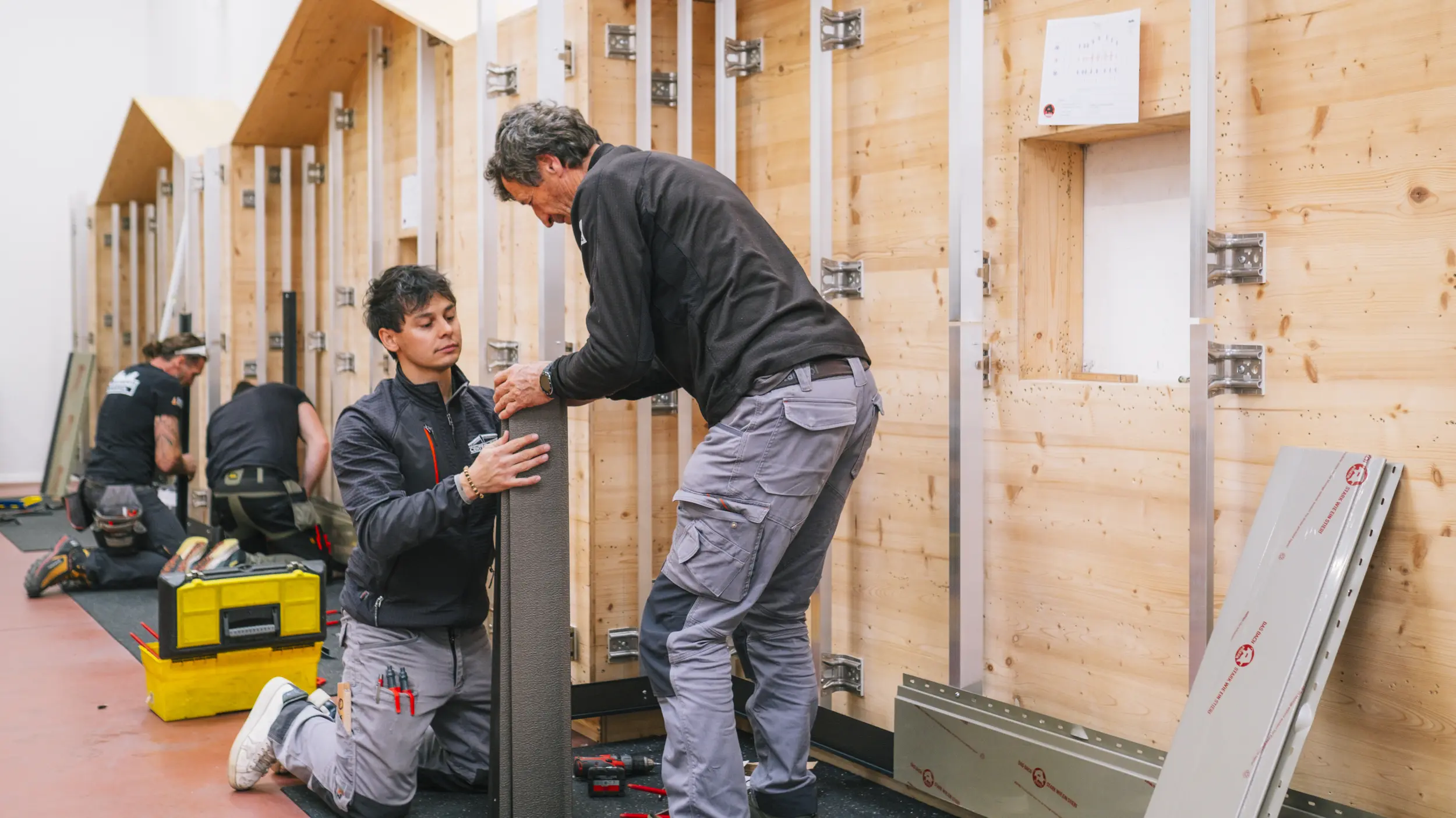 Teilnehmer einer praktischen Schulung in der PREFA Academy Italien arbeiten an einer Holz-Übungswand mit Aluminium-Unterkonstruktion. Zwei Personen im Vordergrund montieren gemeinsam ein Fassadenelement. Die Szene vermittelt handwerkliches Training im Bereich der PREFA Fassaden- und Dachsysteme.