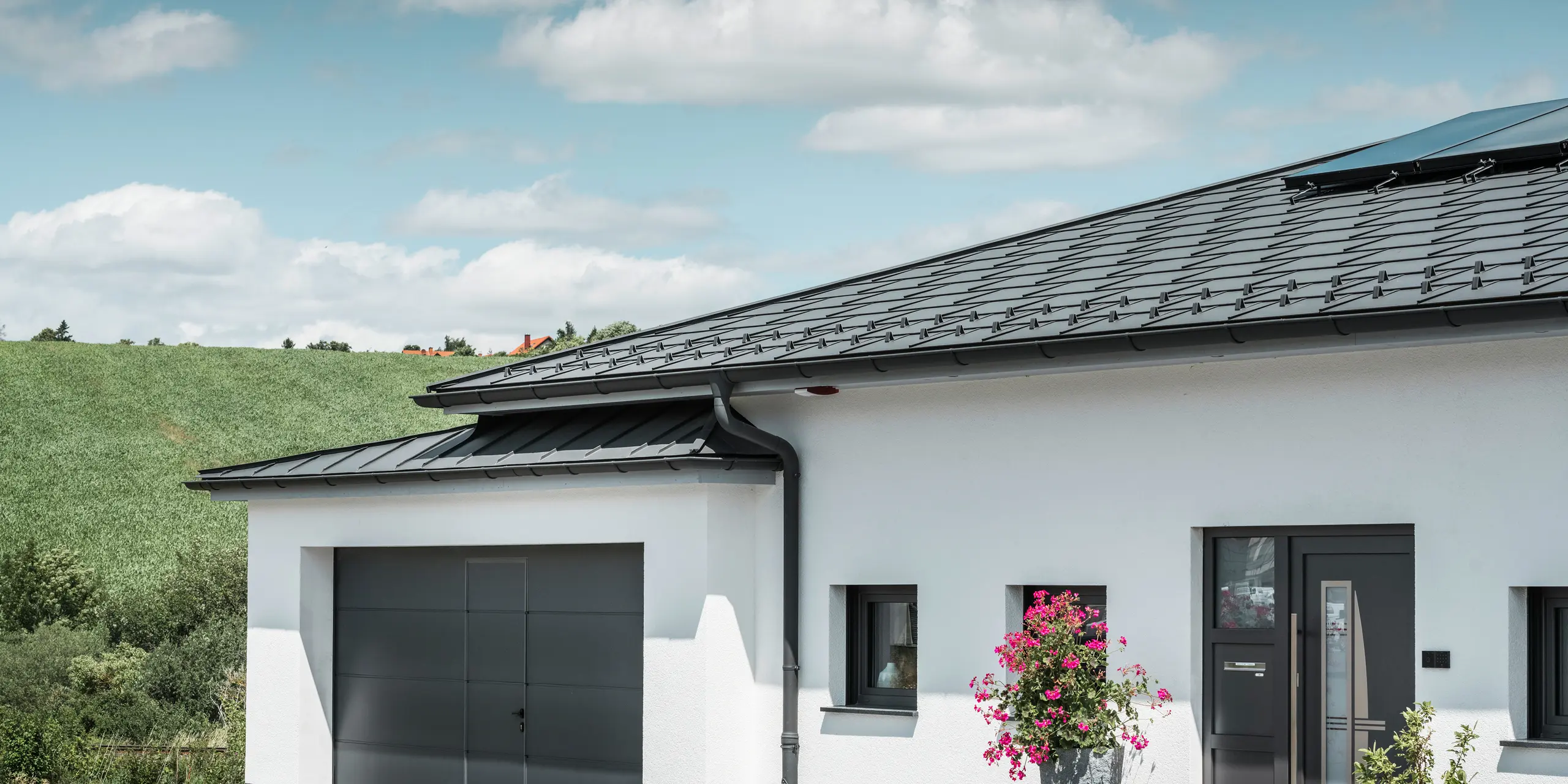 L'immagine mostra la parte anteriore di una casa unifamiliare a Ilmenau, in Turingia, con un elegante tetto PREFA in P.10 grigio scuro. La tegola R.16 conferisce alla casa un aspetto moderno, mentre il sistema di drenaggio integrato PREFA con il canale tondo sospeso e il tubo pluviale grigio scuro garantisce un efficiente deflusso dell'acqua piovana. Il garage, rivestito con PREFALZ grigio scuro, si integra armoniosamente nell'estetica complessiva e sottolinea la qualità e lo stile dei prodotti in alluminio PREFA.