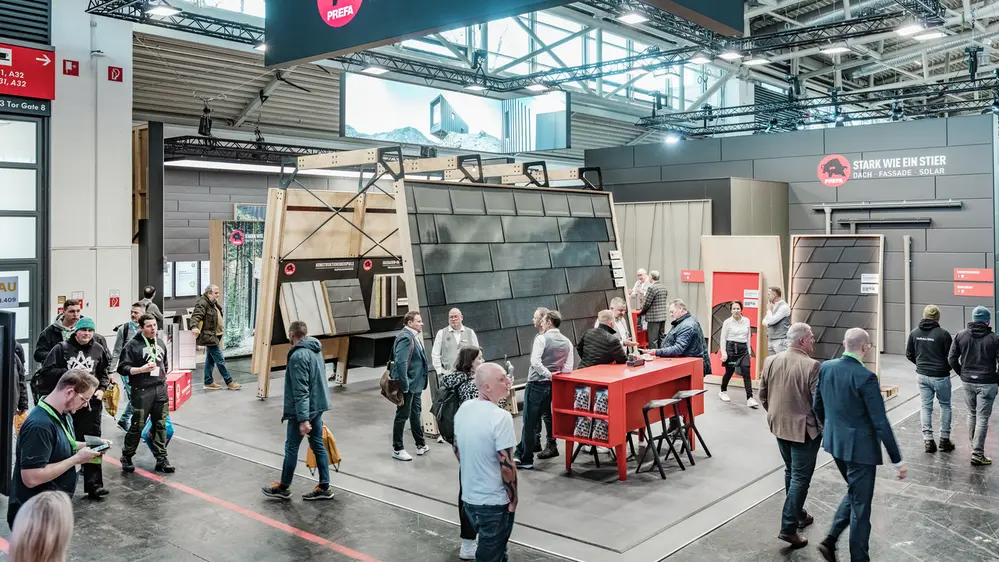 Der PREFA Messestand auf der BAU Messe, gekennzeichnet durch den Slogan „Stark wie ein Stier – Dach, Fassade, Solar“ und eine große hängende Werbefläche. Im Zentrum stehen präsentierte Aluminium-Dach- und Fassadensysteme sowie innovative Montagelösungen. Besucher tauschen sich mit PREFA Mitarbeitern an roten Beratungstischen aus, während weitere Exponate und Infotafeln im Hintergrund zu sehen sind.