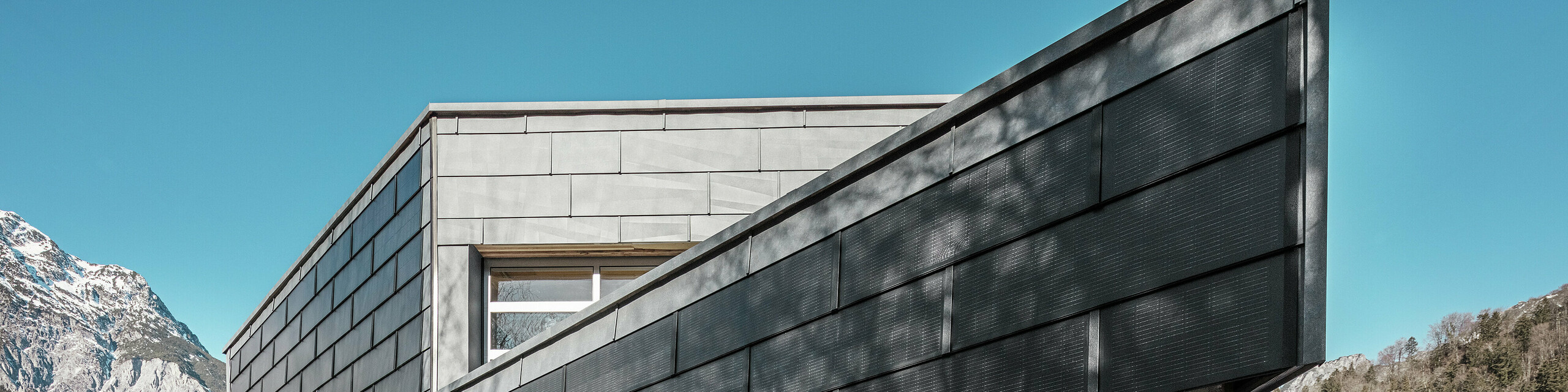 Poslovna zgrada limarske radionice RÜMA u Vorarlbergu impresionira svojom modernom fasadom od PREFA solarnih krovnih ploča i vidljivog betona. Okružena planinama i drvećem čiji se sjena iscrtava na zidu, stoji pod čistim plavim nebom. Oštri kut zgrade i PREFA fasadni paneli FX.12 dodatno naglašavaju upečatljiv izgled.
