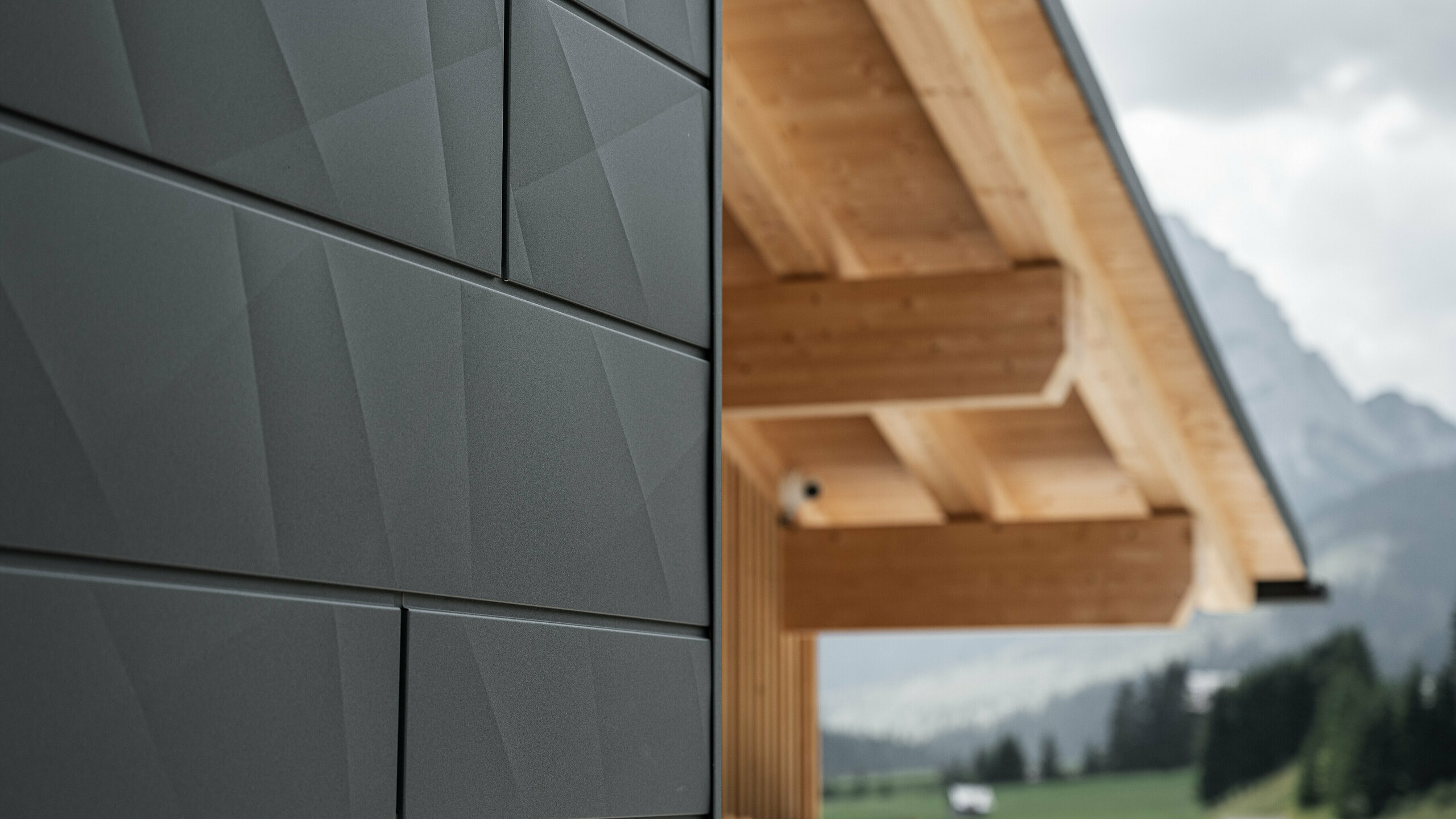 Primo piano dei pannelli di rivestimento Doga.X in P.10 nero di PREFA sulla Casa Hoffe a Sappada, Italia. Il sistema di facciata in alluminio è sapientemente integrato in un design moderno e astratto, che crea un piacevole contrasto con gli elementi in legno a vista della capriata. L'innovativa soluzione per facciate si integra perfettamente nell'ambiente alpino e rappresenta il connubio tra funzionalità, resistenza e cultura edilizia estetica.