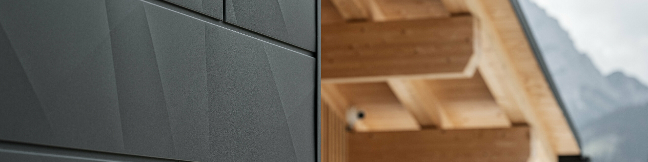 Primo piano dei pannelli di rivestimento Doga.X in P.10 nero di PREFA sulla Casa Hoffe a Sappada, Italia. Il sistema di facciata in alluminio è sapientemente integrato in un design moderno e astratto, che crea un piacevole contrasto con gli elementi in legno a vista della capriata. L'innovativa soluzione per facciate si integra perfettamente nell'ambiente alpino e rappresenta il connubio tra funzionalità, resistenza e cultura edilizia estetica.