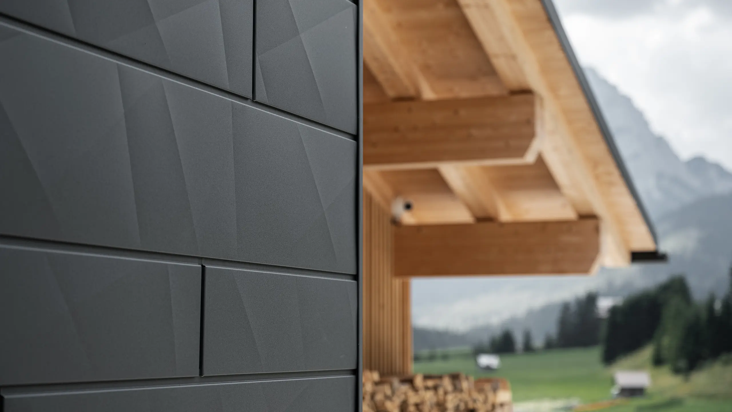 Dettaglio dei pannelli per facciata Doga.X in P.10 nero di PREFA applicati su Casa Hoffe a Sappada (UD). Il sistema di rivestimento in alluminio presenta una posa dal design moderno e astratto, creando un netto contrasto architettonico con le strutture in legno a vista della copertura. La facciata si inserisce armoniosamente nel contesto alpino, coniugando prestazioni tecniche, durabilità e qualità architettonica.