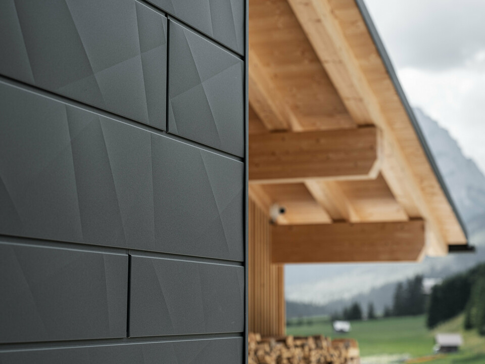 Primo piano dei pannelli di rivestimento Doga.X in P.10 nero di PREFA sulla Casa Hoffe a Sappada, Italia. Il sistema di facciata in alluminio è sapientemente integrato in un design moderno e astratto, che crea un piacevole contrasto con gli elementi in legno a vista della capriata. L'innovativa soluzione per facciate si integra perfettamente nell'ambiente alpino e rappresenta il connubio tra funzionalità, resistenza e cultura edilizia estetica.