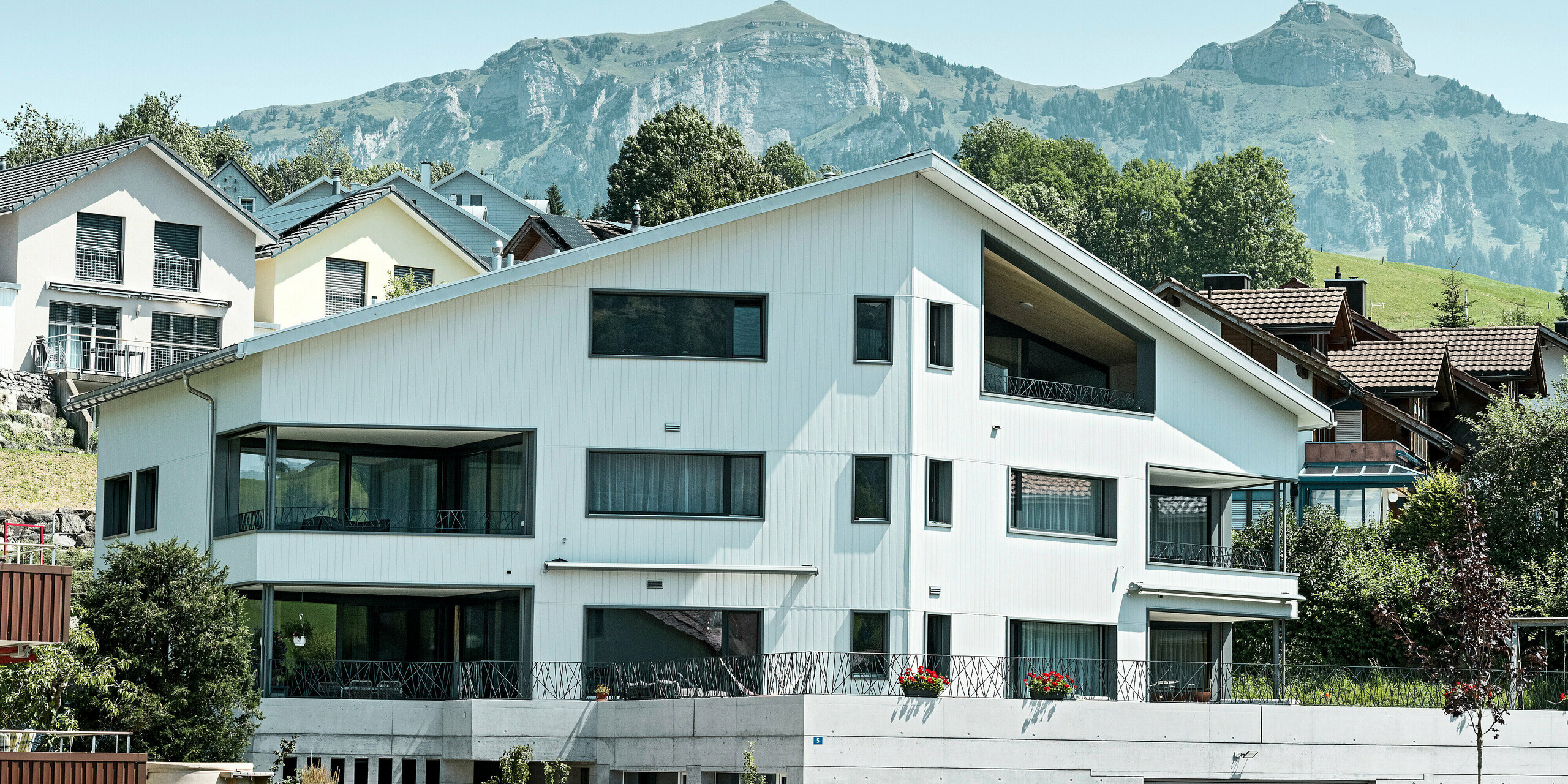 Dieses moderne Mehrfamilienhaus in Weissbad, Schweiz, sticht mit seiner strahlenden PREFA Aluminiumfassade in P.10 Prefaweiß hervor. Die PREFA Sidings sind präzise in vertikaler Ausrichtung angebracht, was dem Gebäude eine klare Struktur verleiht. Die Fassade spiegelt die Flexibilität und Ästhetik wider, für die PREFA Aluminiumprodukte stehen. Die spitze Gebäudeform passt perfekt zu der beeindruckenden Kulisse des majestätischen Alpenpanoramas im Hintergrund.