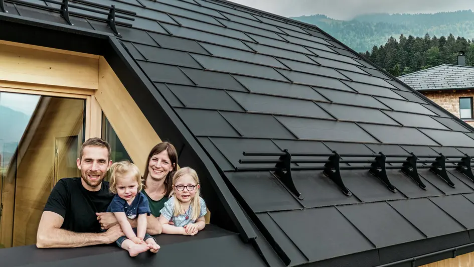 I due committenti con i loro figli si godono la vista dalla loggia della loro casa di nuova costruzione, dotata di eleganti tegole fotovoltaiche PREFA in P.10 nero.