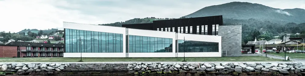 In der Abbildung ist das Healthcare Center in Oystese vom Wasser aus zu sehen. Diese spiegelt sich im Wasser des Hardangerfjord und wurde mit der Wandraute 44x44 in Naturblank eingedeckt. Der Himmel dahinter ist Wolkenbedeckt.