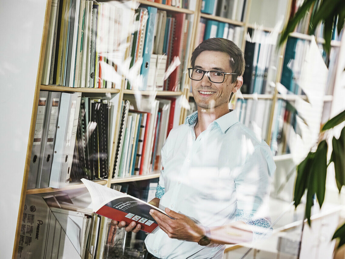 Architekt Christof Künz lächelnd in einem hellen Hemd steht in einem Büro vor einem Bücherregal, das mit Ordnern und Büchern gefüllt ist. Er hält eine geöffnete Broschüre in den Händen. Die Szene wird durch eine Pflanze im Vordergrund und die Spiegelung des Bürointerieurs auf einer Glasscheibe belebt, was eine freundliche und wissensreiche Atmosphäre vermittelt.