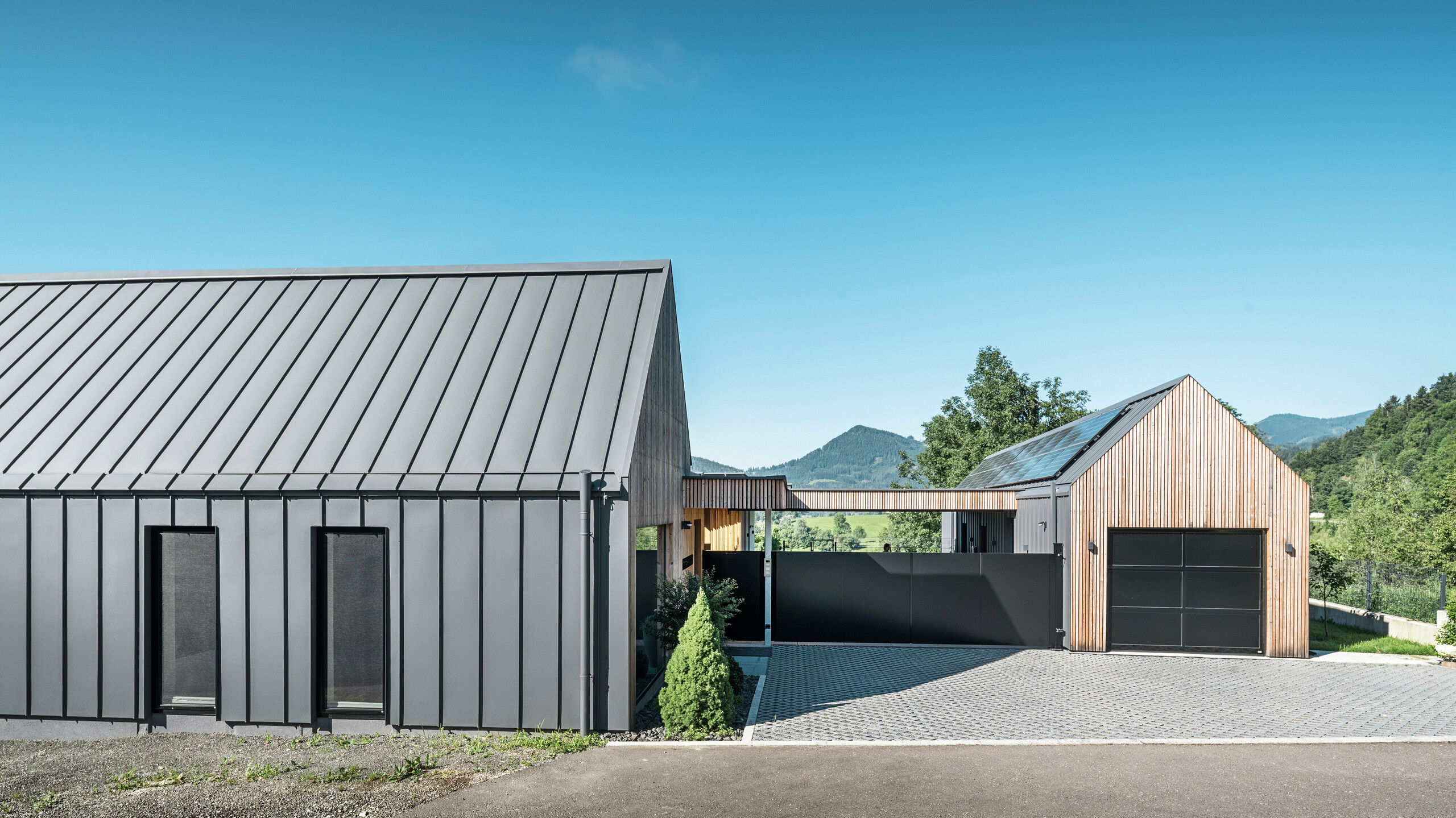 Eingangsbereich eines Einfamilienhauses mit angrenzender Garage in Pogier, Österreich, inmitten einer malerischen Landschaft. Dach und Fassade beider Gebäude sind mit langlebigem PREFALZ in P.10 dunkelgrau verkleidet, was ein modernes Erscheinungsbild erzeugt. Die Garage verfügt über eine harmonisch integrierte Holzverkleidung, die sich nahtlos in die umliegende Landschaft einfügt. Der gepflasterte Hof und die klaren Linien des Entwurfs ergänzen die Ästhetik des Grundstücks, während im Hintergrund die sanften Hügel das Bild einer friedlichen grünen Oase im grünen Herzen Österreichs vervollständigen.