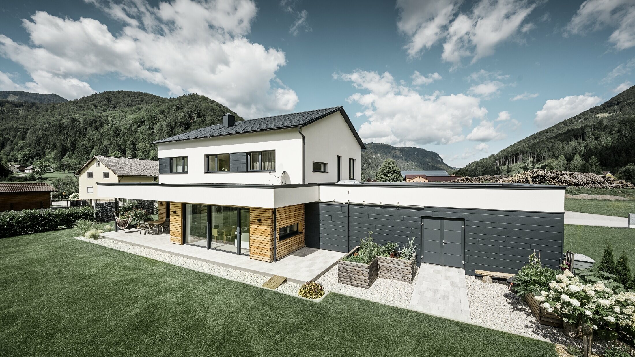 L'immagine mostra una casa unifamiliare scura con intonaco bianco e PREFA Doga.X in P.10 antracite. Il rivestimento scuro della facciata contrasta con la superficie esterna chiara. Le tegole PREFA e il sistema di drenaggio del tetto completano il design durevole. Sullo sfondo colline boscose.