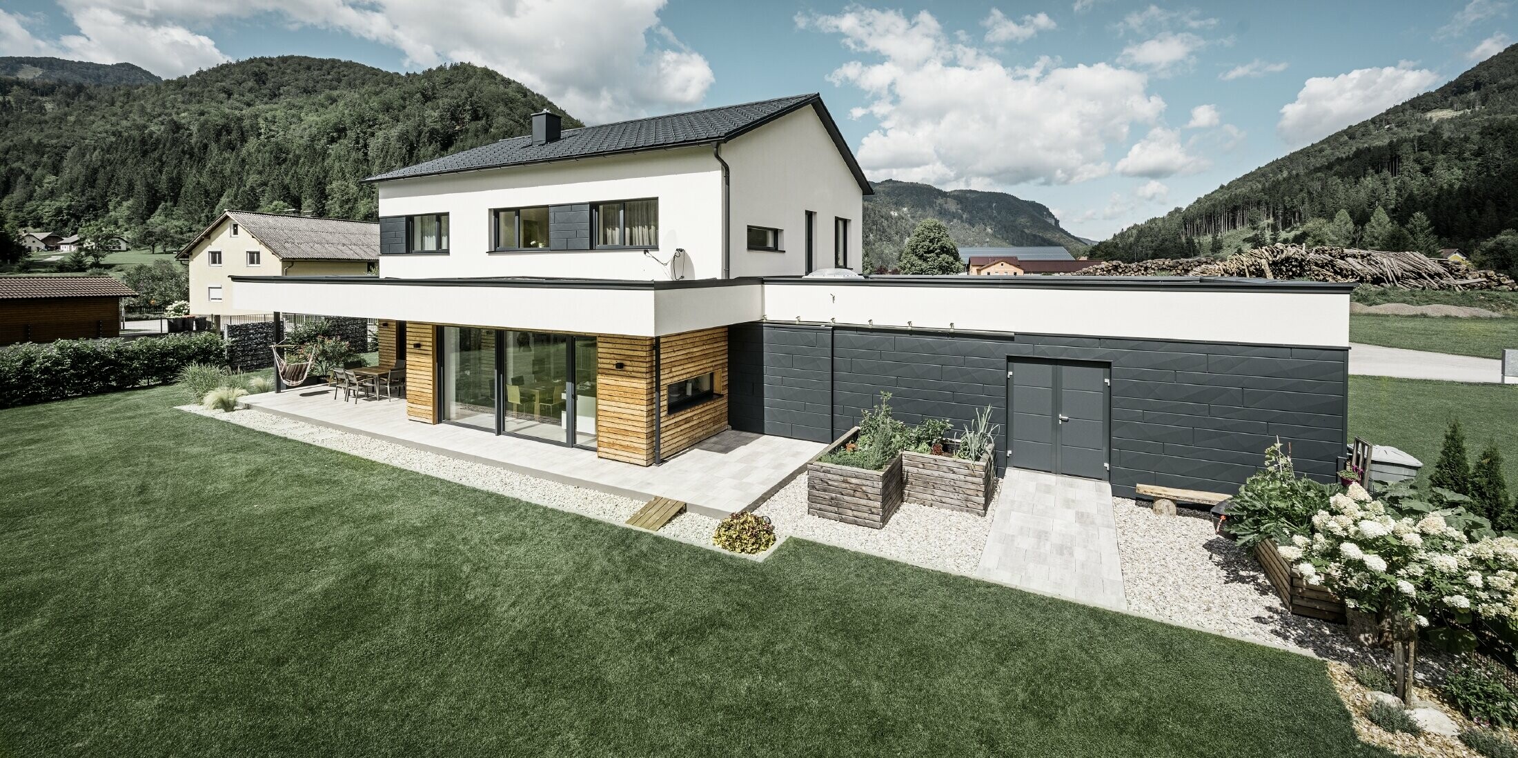 L'immagine mostra una casa unifamiliare scura con intonaco bianco e PREFA Doga.X in P.10 antracite. Il rivestimento scuro della facciata contrasta con la superficie esterna chiara. Le tegole PREFA e il sistema di drenaggio del tetto completano il design durevole. Sullo sfondo colline boscose.