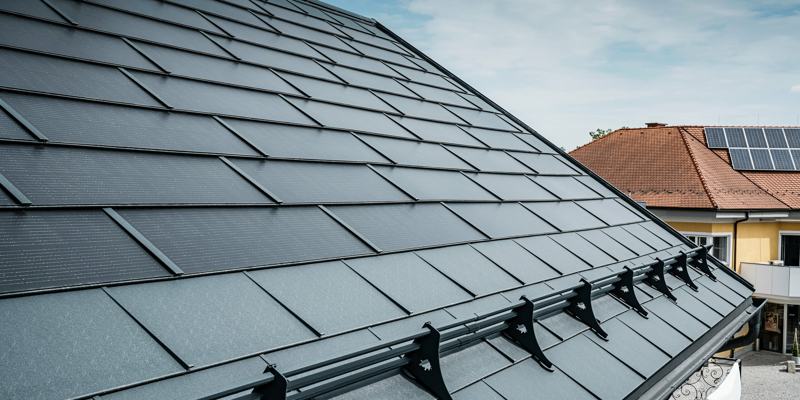 Vista di un tetto moderno con tegole fotovoltaiche PREFA color antracite. Vicino alla gronda, il tetto è stato dotato delle classiche tegole R.16 PREFA e di un sistema paraneve di PREFA. Sullo sfondo si vede il tetto di una casa vicina con tegole tradizionali e pannelli fotovoltaici sul tetto, che evidenzia il contrasto tra il design del tetto nuovo e quello tradizionale. L'innovativa tegola fotovoltaica PREFA combina estetica e funzionalità e offre una soluzione energetica sostenibile per tutti i tipi di edifici.