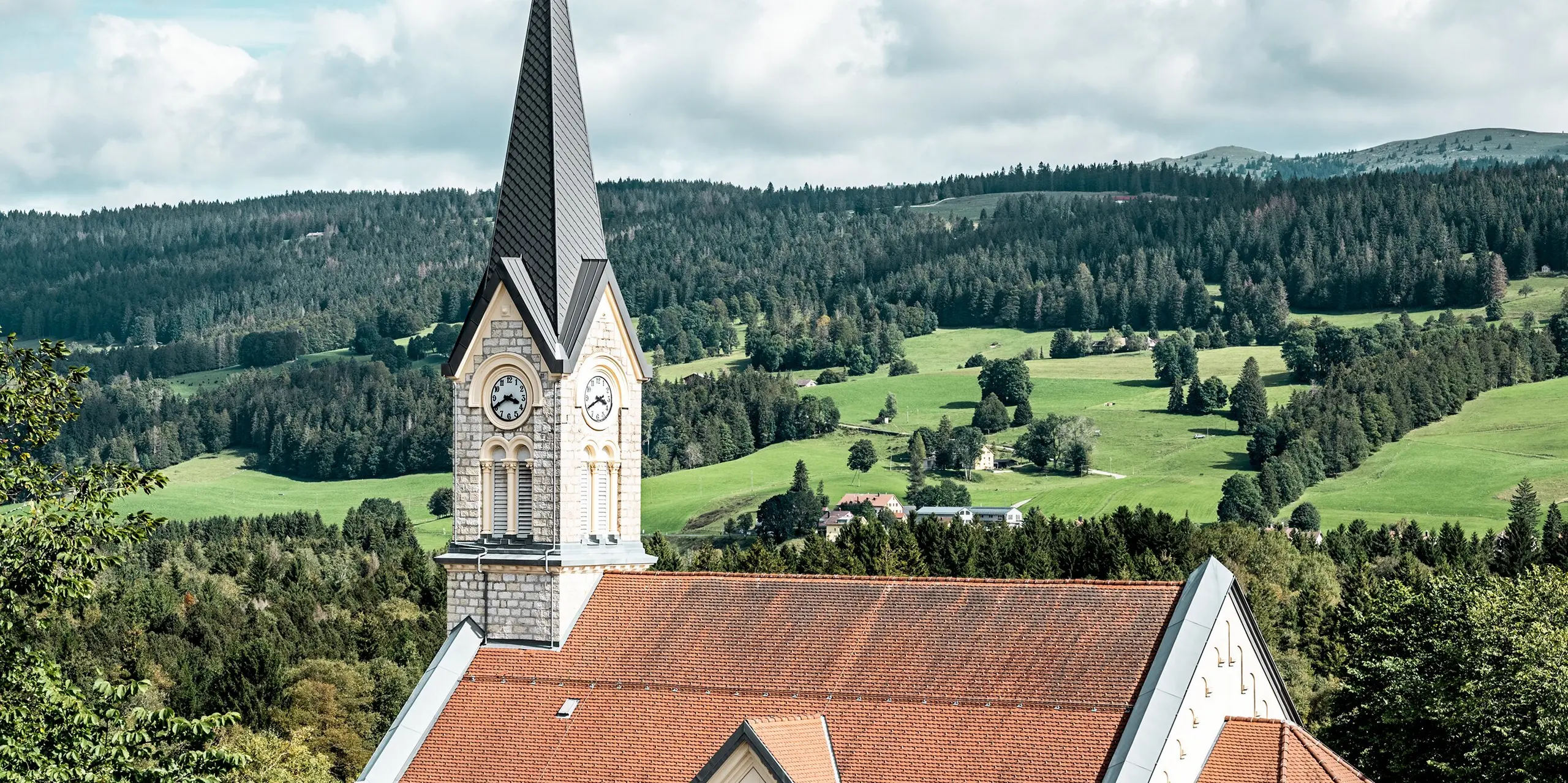 Kirche in Le Sentier vor der beeindruckenden Hügellandschaft des Jura-Gebirges. Die schlanke Turmspitze mit PREFA Dachraute 29 × 29 in P.10 Dunkelgrau setzt einen markanten architektonischen Akzent. Die hochwertige Aluminiumdeckung überzeugt bei steilen Dachformen durch Langlebigkeit, Witterungsbeständigkeit und eine präzise Optik, die historische Bausubstanz modern ergänzt.