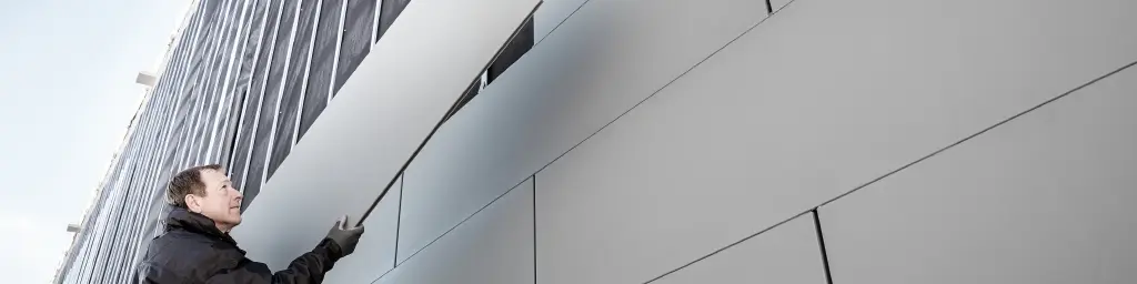 Immagini di un cantiere in corso d'opera con rivestimento in Doga PREFA formato 500. L'installatore procede al fissaggio di un pannello di facciata. Ripresa dal basso. 