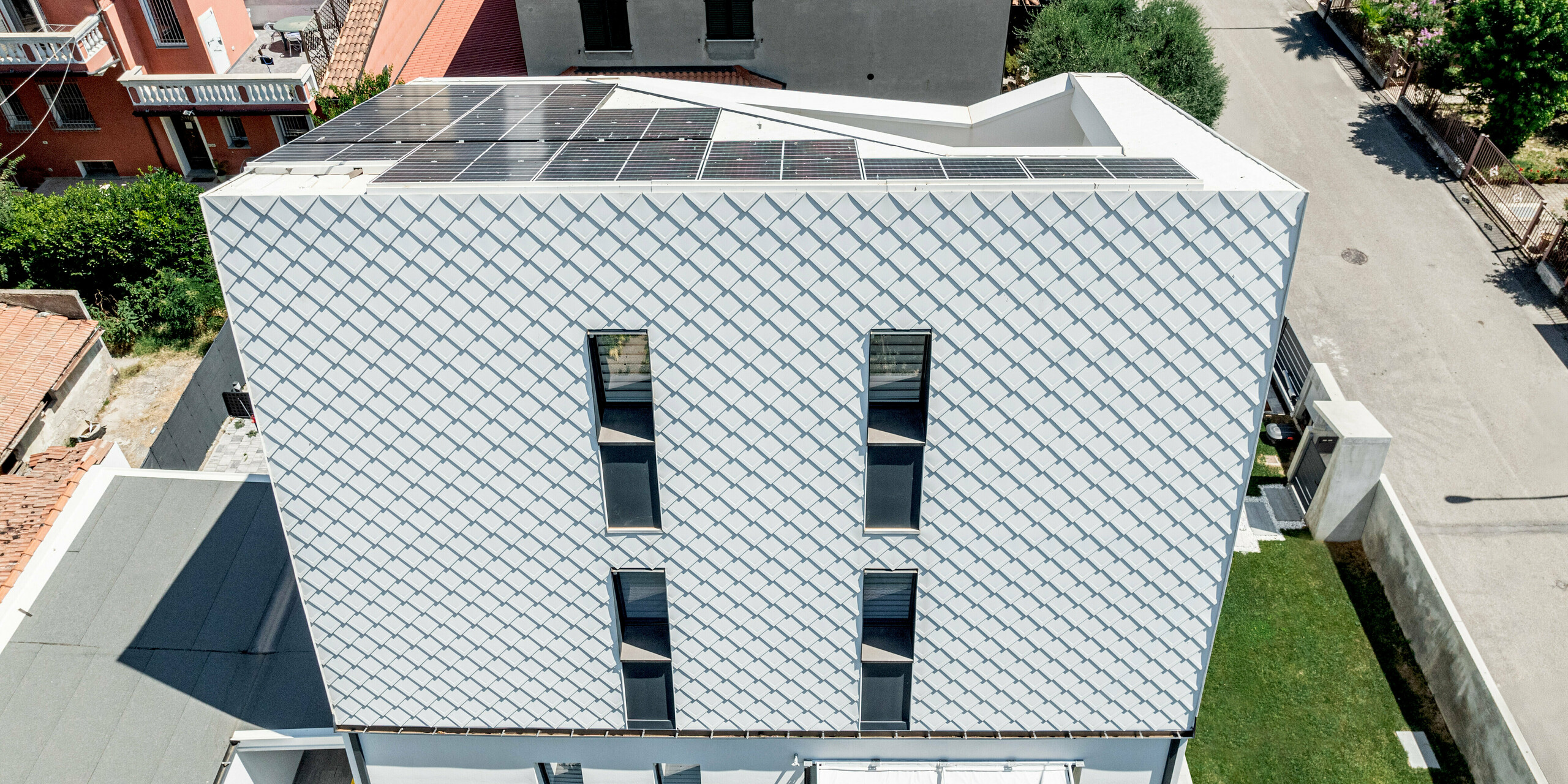 Luftaufnahme eines modernen Einfamilienhauses in Forlì, Italien, dessen markante Fassade mit PREFA Wandraute 29 × 29 in P.10 Reinweiß sofort ins Auge fällt. Die Aluminiumrauten erzeugen eine lebendige Struktur und spannende Licht- und Schattenspiele auf der geneigten Fläche. Umgeben von traditionellen Ziegeldächern hebt sich die strahlend weiße PREFA Aluminiumfassade deutlich ab und überzeugt zugleich durch Langlebigkeit, Witterungsbeständigkeit und nahezu wartungsfreie Qualität.