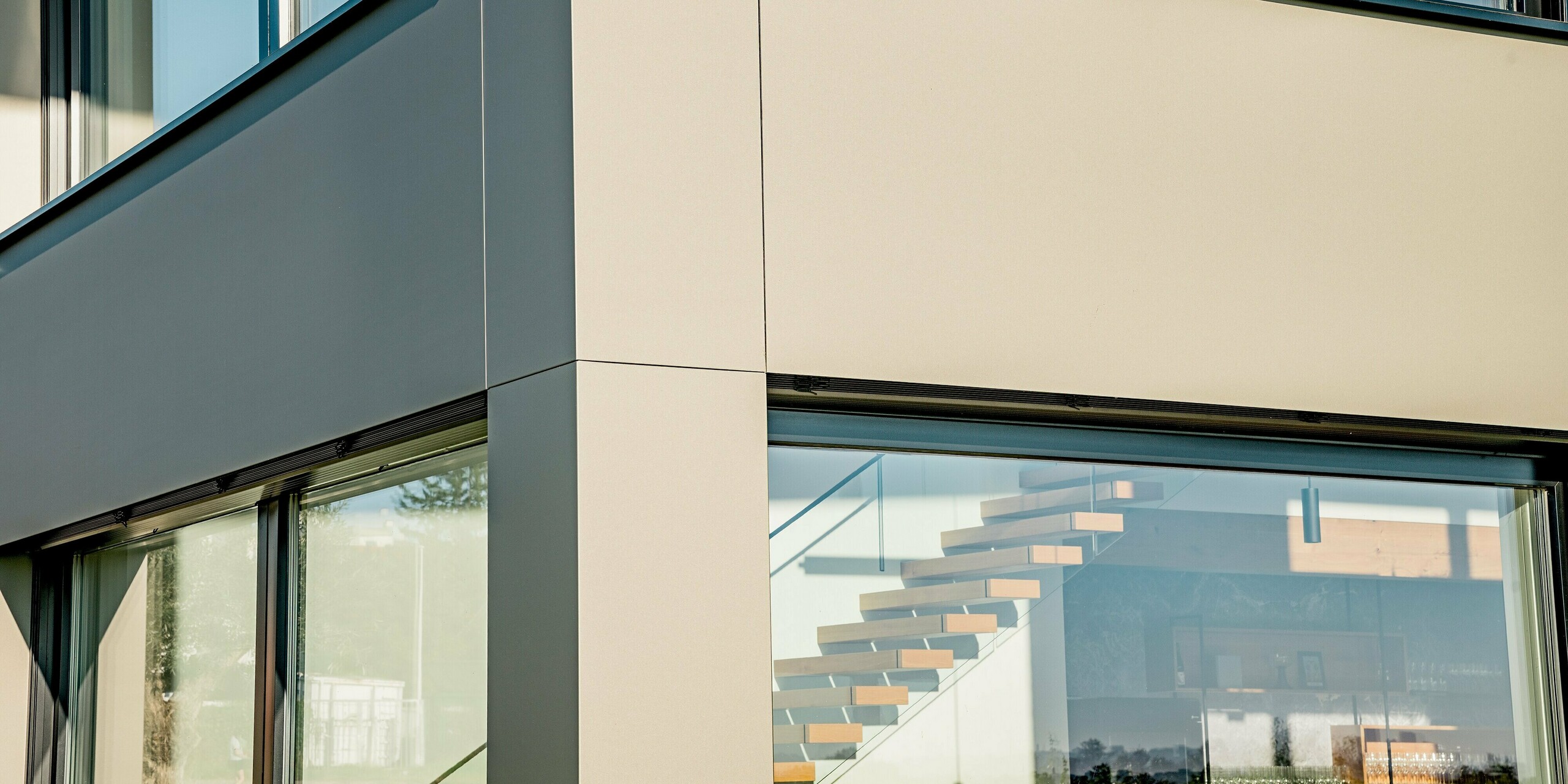 Detailaufnahme der Gebäudeecke des Weinguts Nigl, die mit PREFA Aluminium Verbundplatten in der Farbe P.10 Prefabronze verkleidet ist. Die klar erkennbaren, präzise ausgeführten Fugen betonen den eleganten Kontrast zu den Glasflächen und unterstreichen die moderne, hochwertige Architektur. Die metallische Oberfläche wirkt glatt, edel und zugleich robust. Durch die großzügige Glasfassade ist die stilvolle, freischwebende Holztreppe im Inneren sichtbar, was für ein spannendes Spiel zwischen Außen- und Innenraum sorgt. Die PREFABOND-Platten bieten dauerhaften Witterungsschutz, sind wartungsfrei und langlebig – eine ideale Lösung für designorientierte Gewerbebauten, die Qualität und Beständigkeit verlangen.