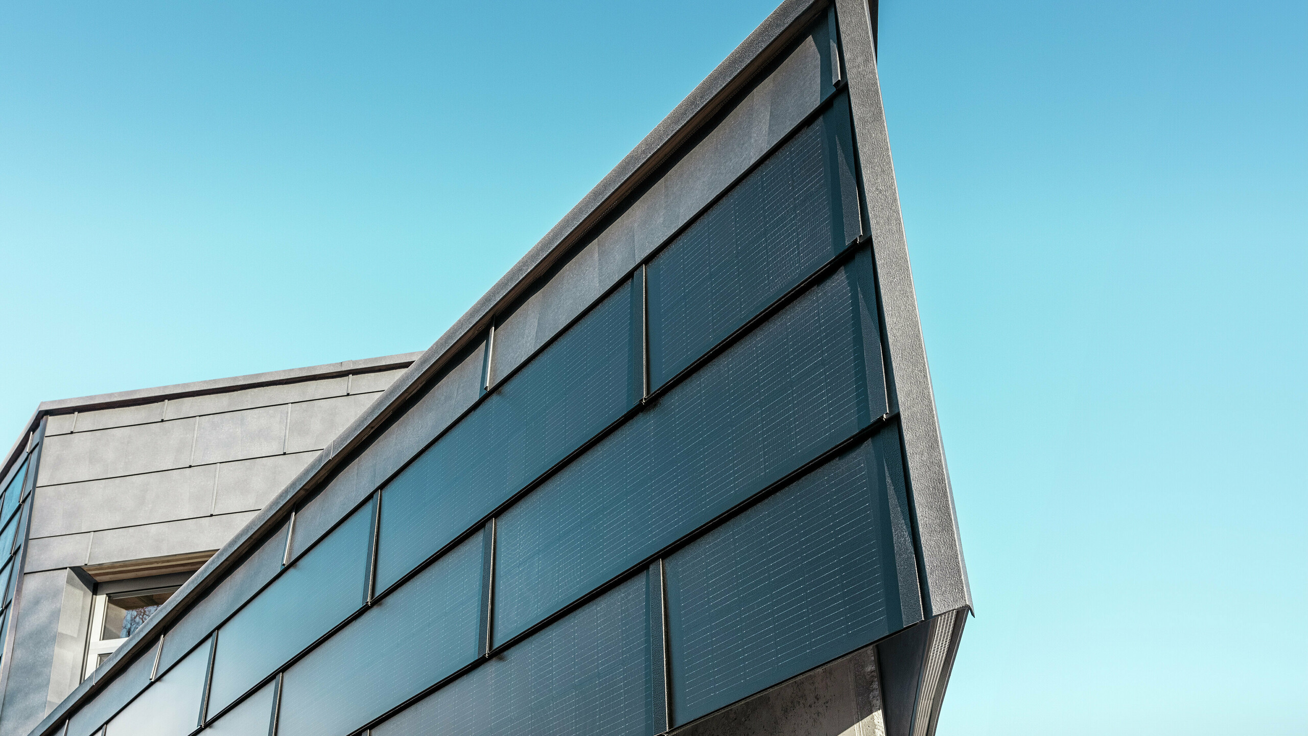 Moderna poslovna fasada limarske radionice RÜMA u Vorarlbergu s PREFA solarnim krovnim pločama od aluminija i integriranim fotonaponskim modulima, u kombinaciji s PREFA fasadnim panelima FX.12, za održivu proizvodnju energije i dinamičan dizajn fasade. Snimka prikazuje kut zgrade izbliza. U pozadini se vidi vedro plavo nebo.