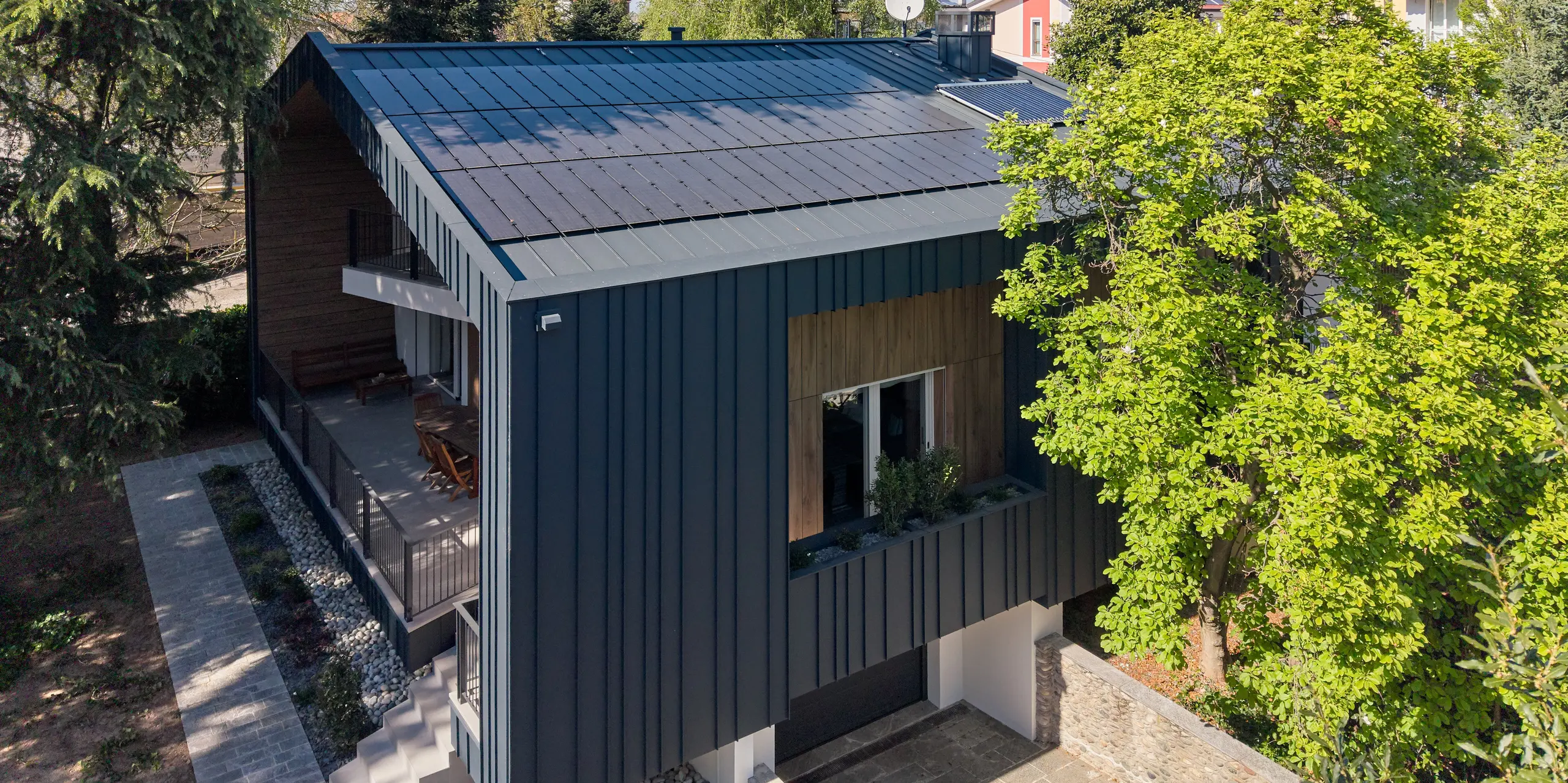 Detailansicht der Villa Castellanza mit durchgängiger PREFALZ Dach- und Fassadengestaltung in P.10 Anthrazit. Großflächig integrierte PREFALZ Solarmodule prägen die moderne Gebäudehülle und schaffen ein harmonisches Gesamtbild. Die langlebigen PREFA Aluminiumprodukte verbinden elegante Architektur, nachhaltige Energiegewinnung und hohe Widerstandsfähigkeit. Umgeben von Bäumen wirkt das Wohnhaus besonders eindrucksvoll.
