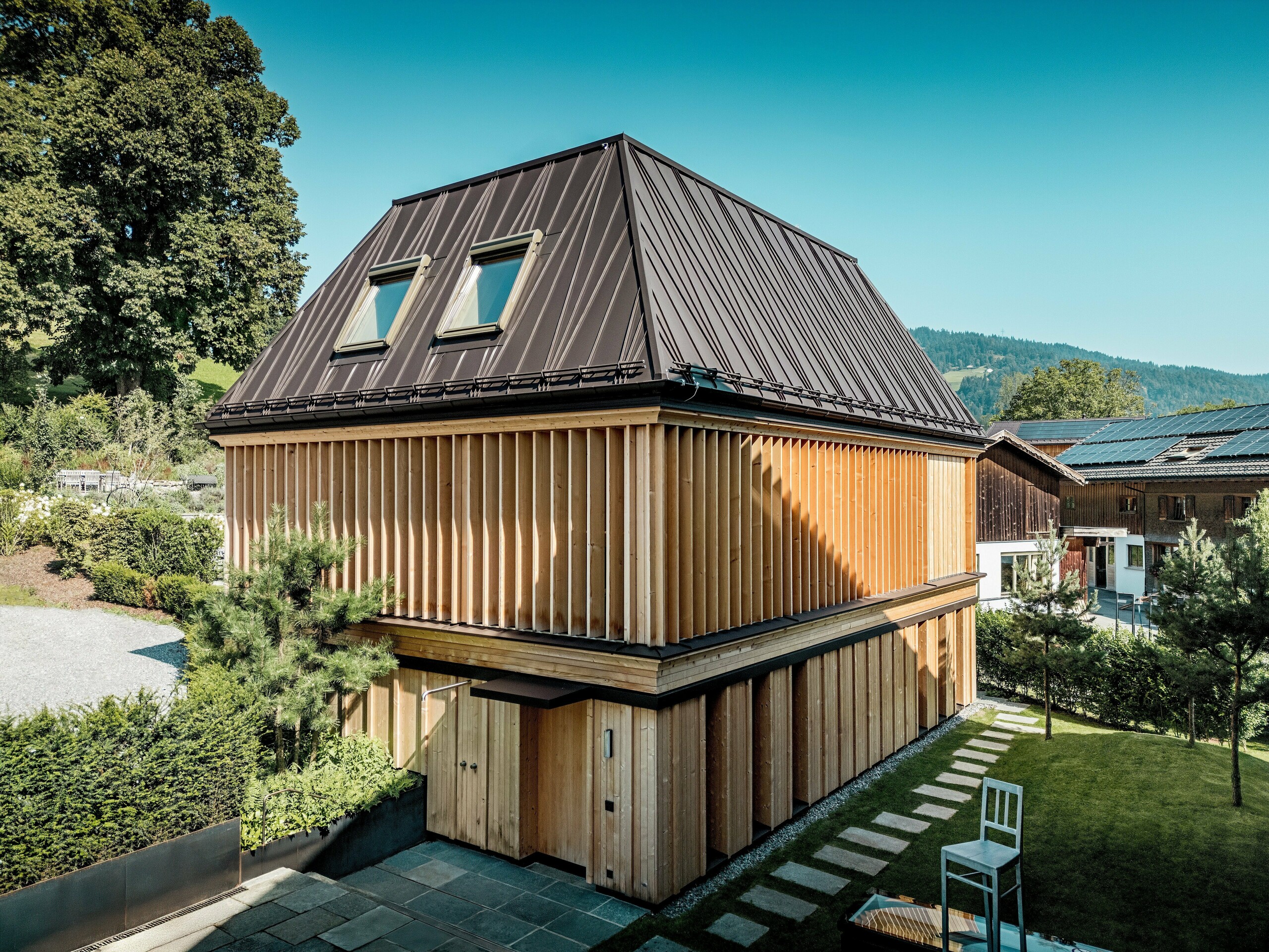 Das „Badehaus“ des Hotel Hirschen in Schwarzenberg in einem klaren, natürlichen Licht. Im Fokus steht das außergewöhnliche PREFA Stehfalzdach in P.10 Nussbraun mit seiner präzisen Ausführung und den markanten, teils diagonal verlaufenden Stehfalzen. Zwei Dachfenster sorgen für natürliches Licht im Inneren. Die Fassade ist mit geschoßhohen, vertikal angeordneten Holzlamellen gestaltet, die sowohl gestalterisch als auch funktional zur Geltung kommen. Im Vordergrund führen Natursteinplatten durch einen gepflegten Gartenbereich. Umgeben von traditionellen Holzhäusern und sanften Hügeln fügt sich das moderne Gebäude harmonisch in das Ortsbild des Bregenzerwaldes ein.
