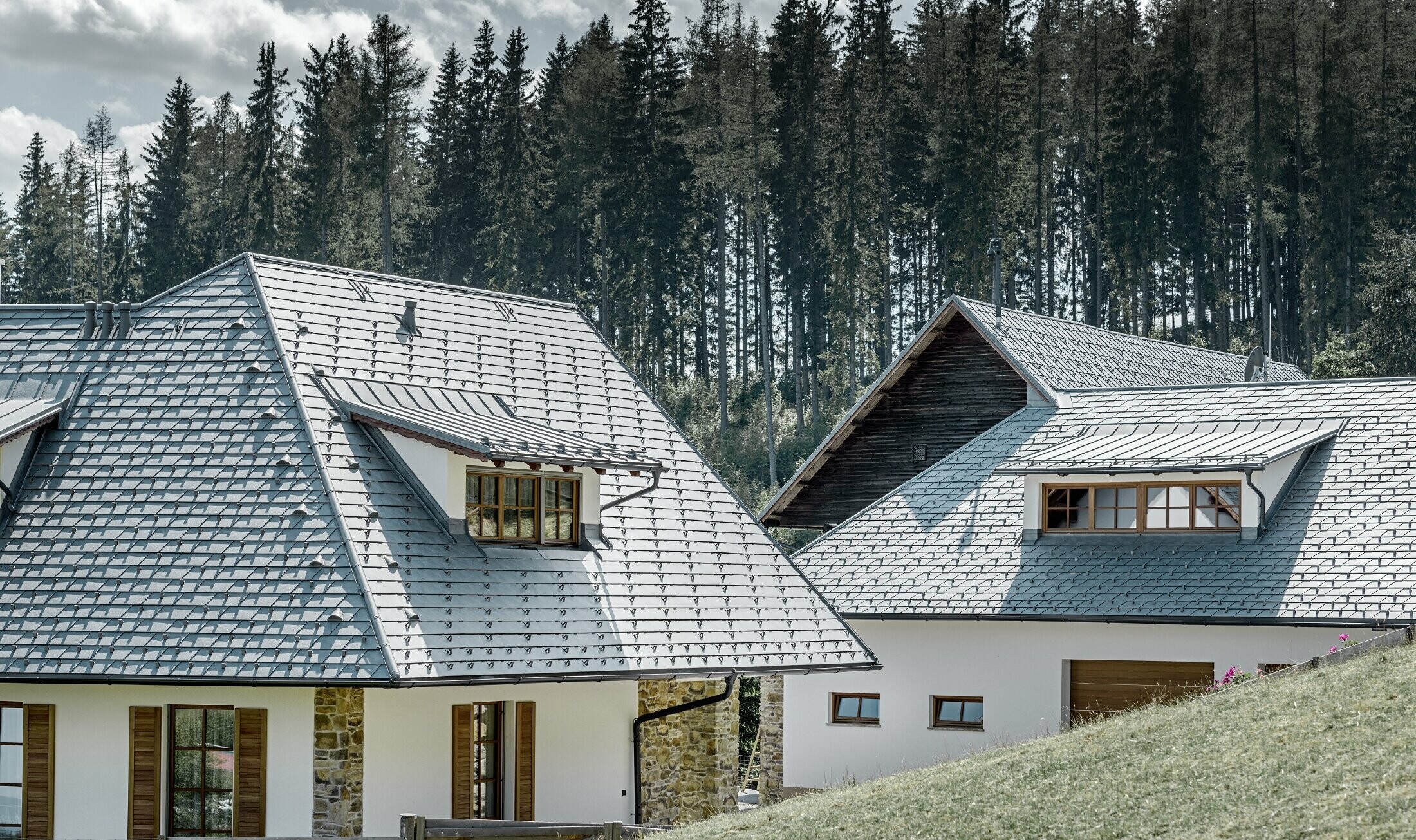 Veduta posteriore della casa unifamiliare in pendenza; il tetto è stato coperto con la scandola in alluminio color grigio pietra, gli abbaini con lamiera con aggraffatura, sempre in grigio pietra. Sullo sfondo si staglia un bosco.