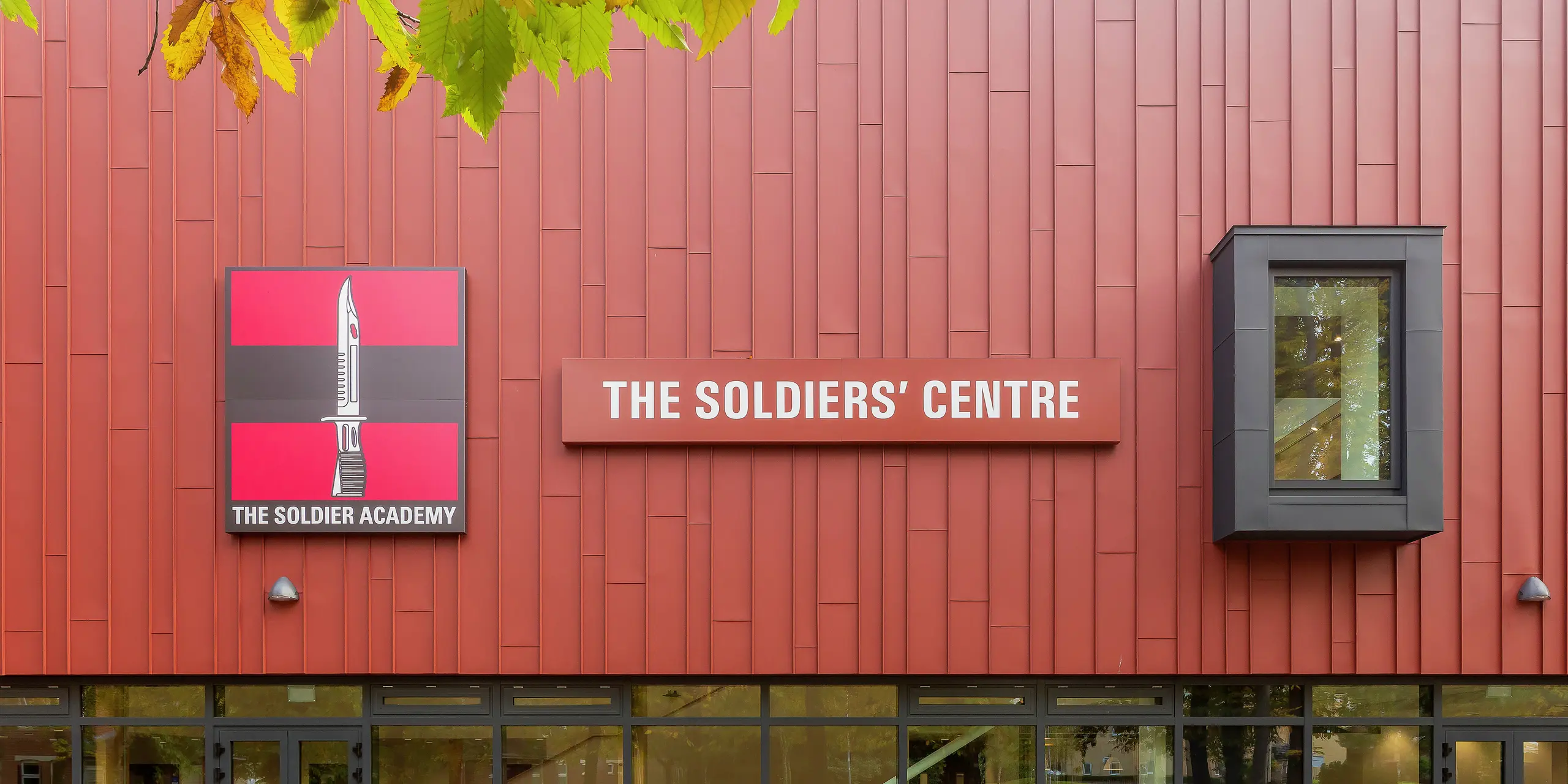 Speziell für die Armee errichtete Sozialeinrichtung mit PREFALZ-Fassade: Die präzise verarbeitete Stehfalzdeckung in P.10 Oxydrot verleiht dem Gebäude ein markantes Erscheinungsbild. Der Eingangsbereich besticht durch großzügige Glasfronten, die für Helligkeit und Transparenz sorgen. Das Logo der 'Soldier Academy' und das auffällige Gebäudeschild setzen klare optische Akzente. Die Fensterrahmen in P.10 Anthrazit runden das Design harmonisch ab. Hochwertige PREFA Aluminiumprodukte für langlebige und ästhetische Architektur.