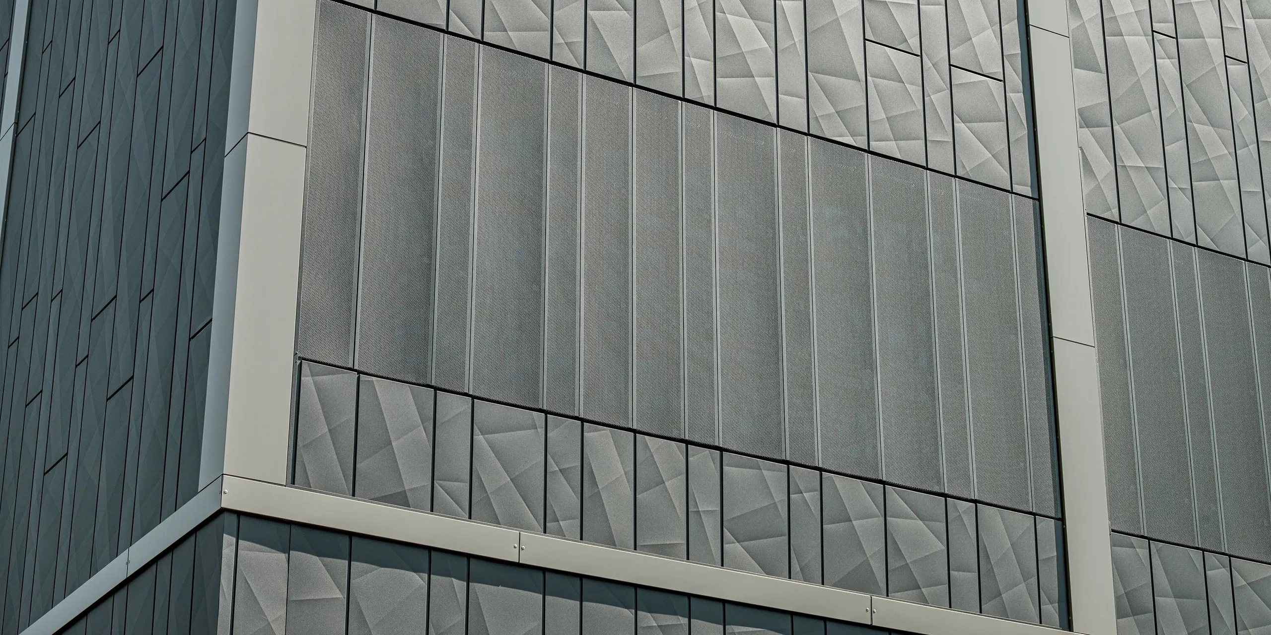 Das Bild zeigt einen detailreichen Ausschnitt der Aluminiumfassade des Einkaufszentrums in Rohrbach. Besonders auffällig sind die anthrazitfarbenen Siding.X-Paneele mit ihrer geometrischen Struktur und den variierenden Oberflächen, die einen dynamischen, modernen Eindruck vermitteln.  Im unteren Bereich des Bildes sind perforierte Siding-Elemente zu sehen. Diese setzen nicht nur ästhetische Akzente, sondern sorgen auch für eine gezielte Belüftung und Lichtdurchlässigkeit. Die Kombination aus glatten und perforierten Paneelen verleiht der Fassade somit eine lebendige und funktionale Gestaltung, die optisch und praktisch überzeugt.