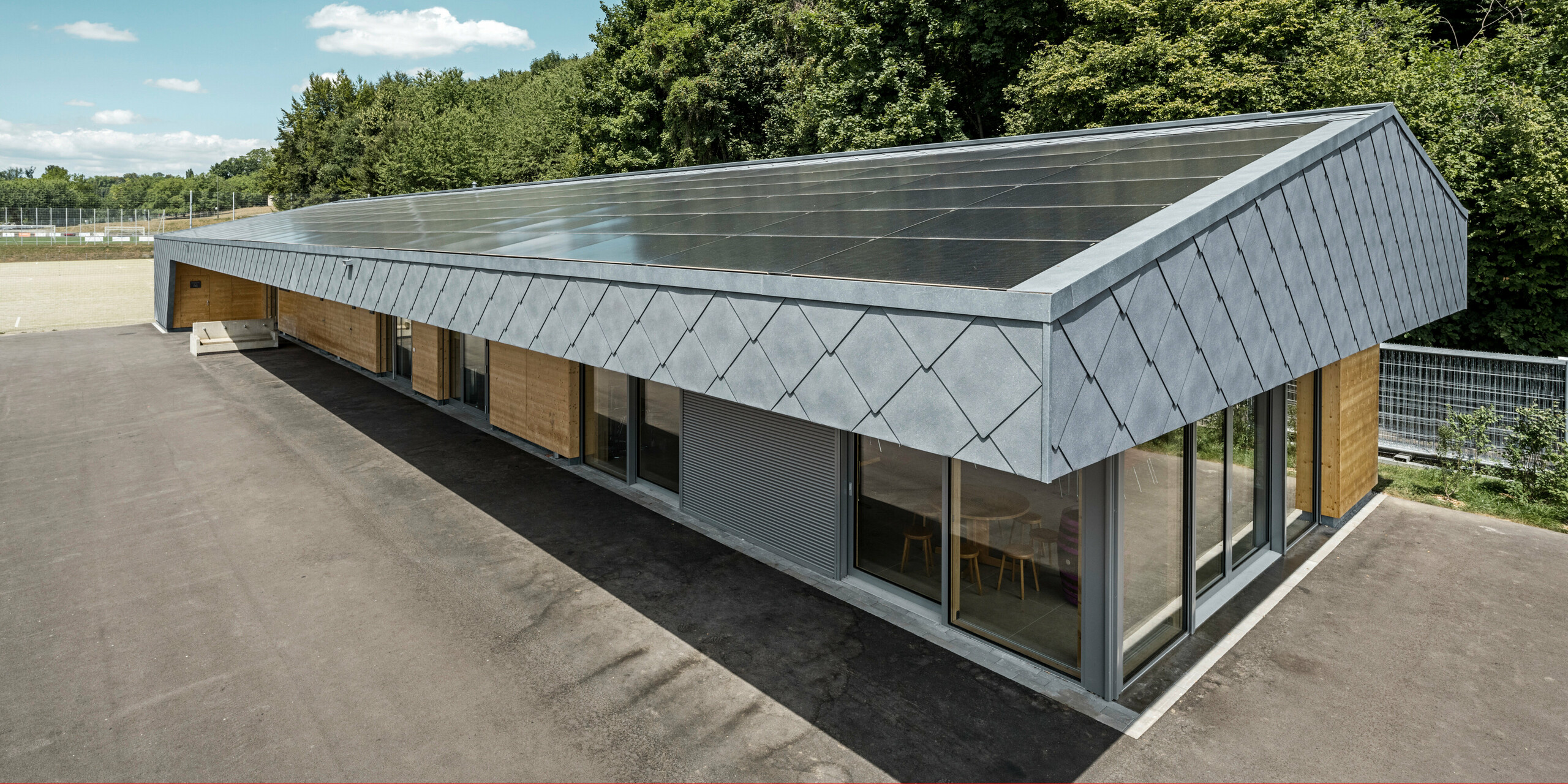 Ansicht von oben auf eine moderne Sportanlage in Prilly, Schweiz. Das Gebäude wurde mit mit langlebigen PREFA Dach- und Wandrauten 44x44 in der Farbe P.10 Steingrau ausgestattet. Die Rauten aus Aluminium sorgen für ein markantes Design, das perfekt mit der großflächig installierten Photovoltaikanlage harmoniert. Diese Kombination zeigt die innovative Verbindung von Funktionalität, Energieeffizienz und ansprechender Ästhetik, die für PREFA Aluminiumprodukte steht. Das Gebäude repräsentiert zukunftsorientiertes Bauen mit einem Fokus auf Nachhaltigkeit und Energieautonomie.