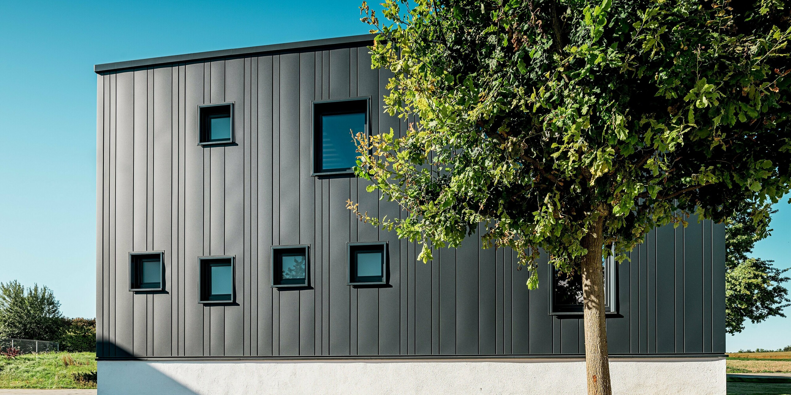 Die Seitenansicht des Firmengebäudes in Neckarsulm zeigt die harmonische Fassadengestaltung mit verschieden breiten PREFA Sidings, die eine moderne, rhythmische Struktur erzeugen. Die kompakte Gebäudeform wird durch die dunkle Farbgebung in P.10 Anthrazit zusätzlich betont. Die robuste Aluminiumfassade bewahrt langfristig ihr Erscheinungsbild und schützt zuverlässig vor äußeren Einflüssen. Die Natur im Vordergrund verstärkt den architektonischen Kontrast.