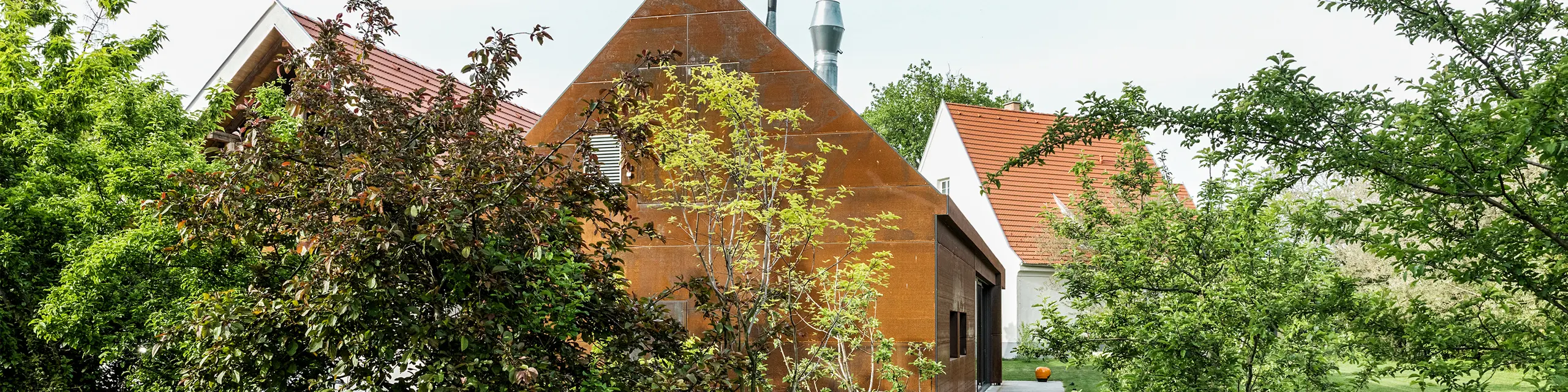 Residenza Velich nella regione del Seewinkel in Burgenland: Vista del moderno edificio, circondato da una rigogliosa vegetazione. La residenza funge da sede per il dialogo di esperti delle PREFARENZEN 2025, durante il quale architetti ed esperti di PREFA selezionano i più interessanti oggetti architettonici. In primo piano si vede una elegante parete metallica color ruggine con la scritta 'RESIDENZ VELICH'. L'architettura dell'edificio combina elementi tradizionali e moderni e si integra armoniosamente con l'ambiente naturale.